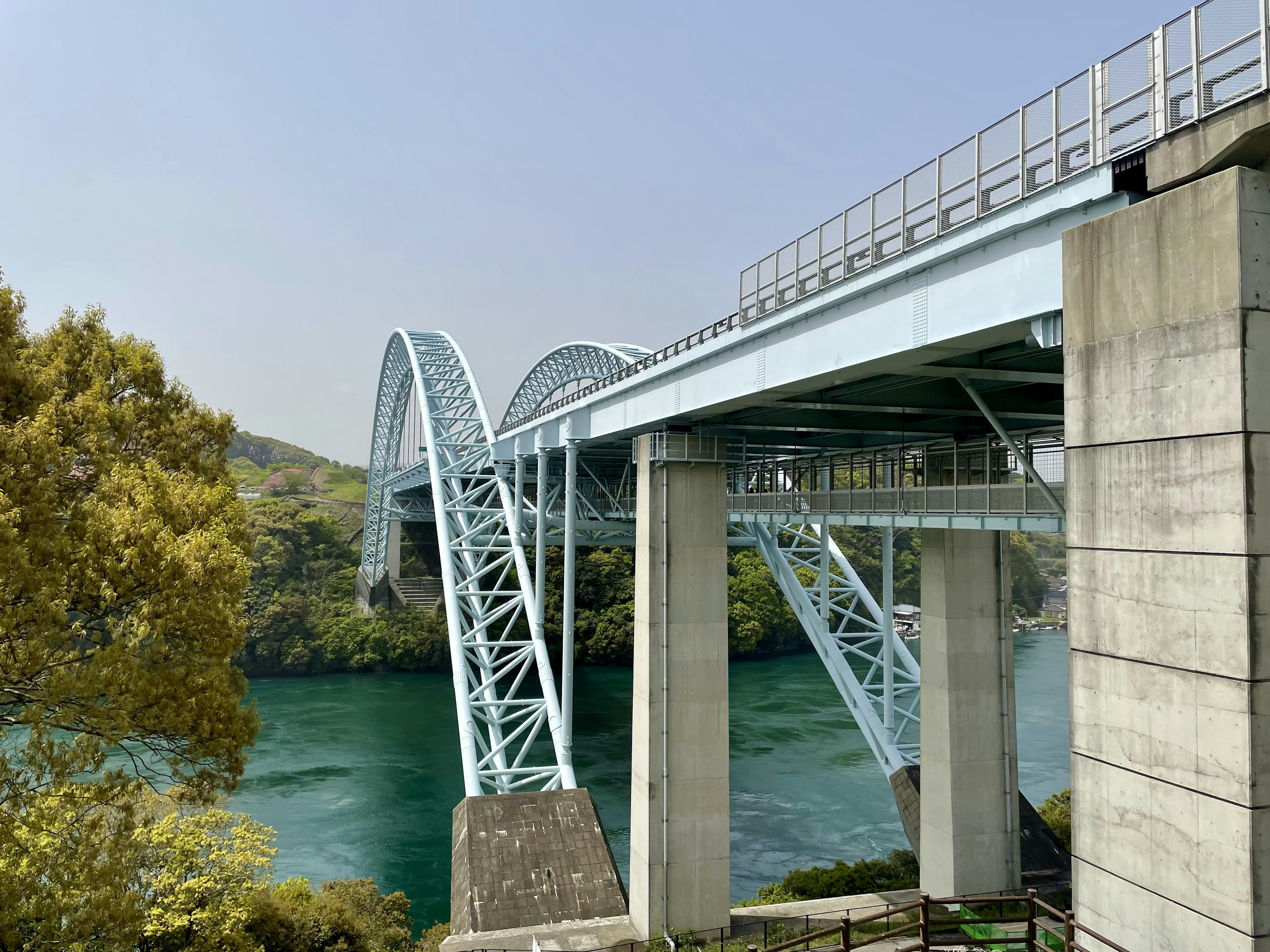 青い橋が川を跨いでいる風景 橋の下にコンクリートの支柱 周囲に緑の木々が見える