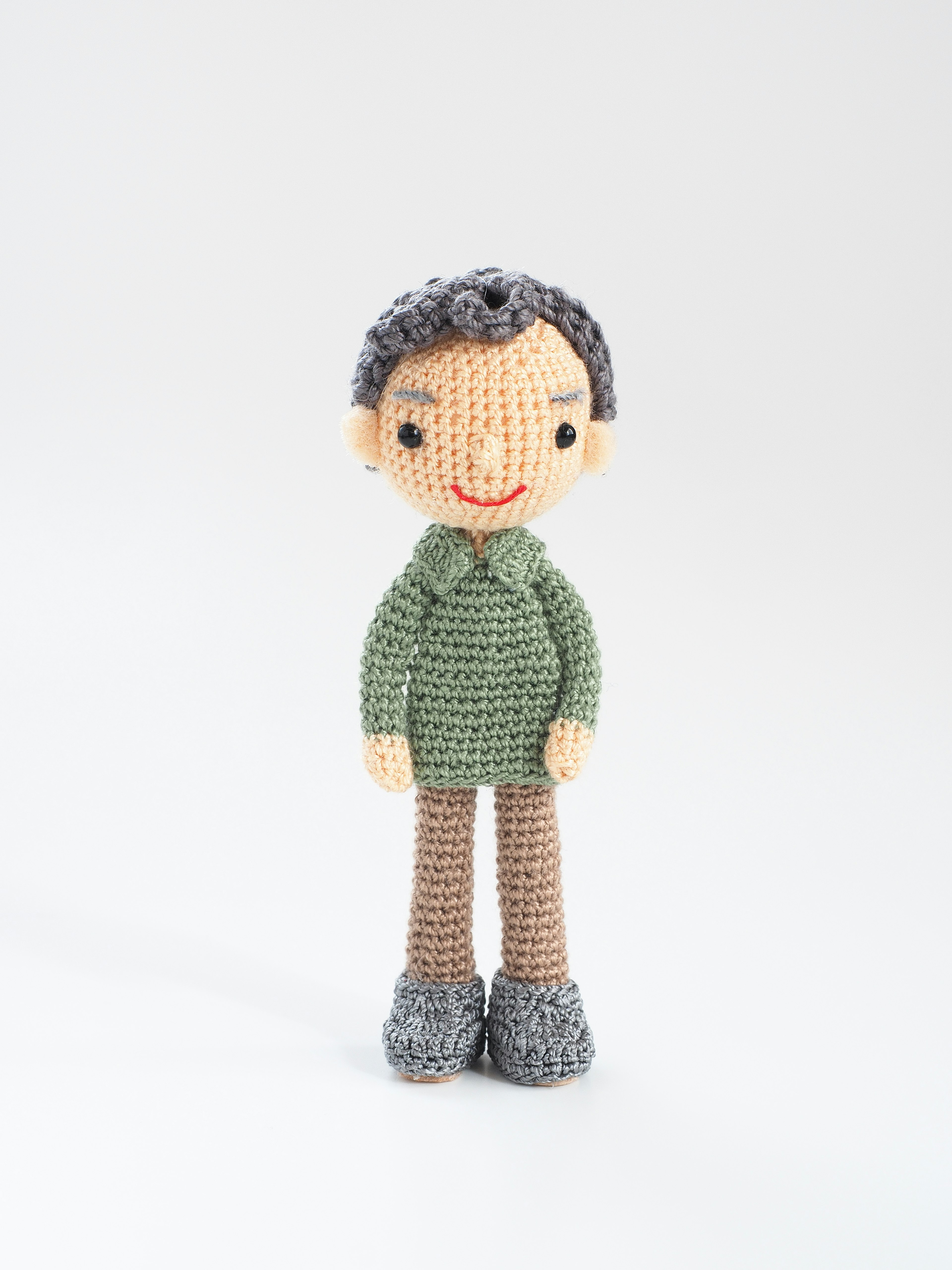 A crochet doll of a boy standing wearing a green sweater and brown pants