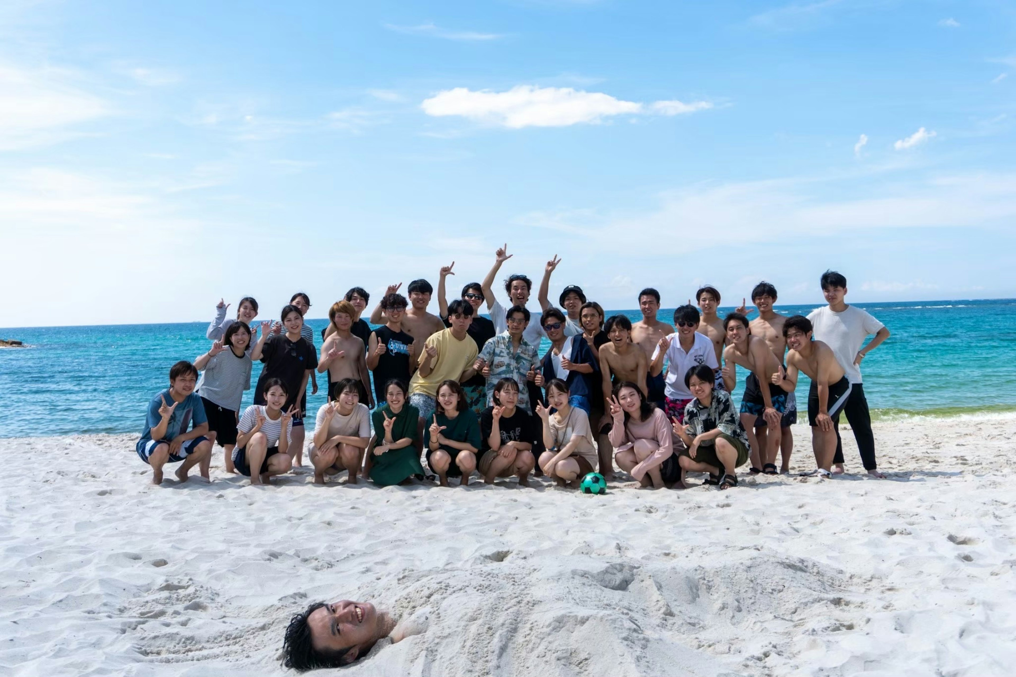 Gruppenfoto am Strand mit blauem Himmel und Meer, Mann im Sand begraben