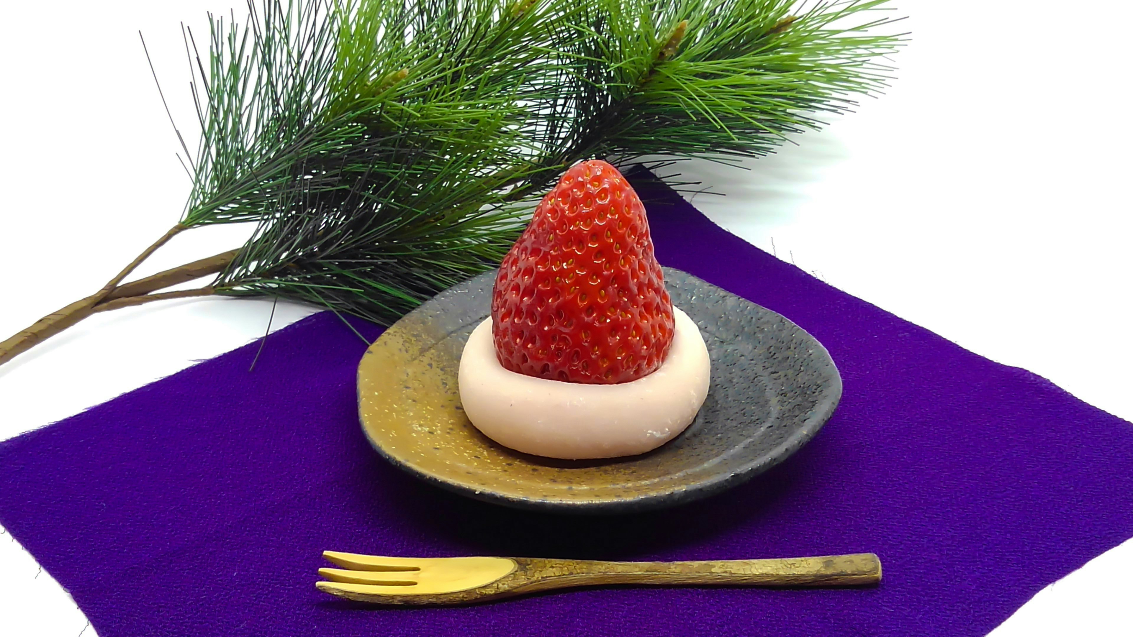 Un dessert avec une fraise sur un mochi rond dans une assiette noire avec une fourchette et des feuilles de pin en arrière-plan