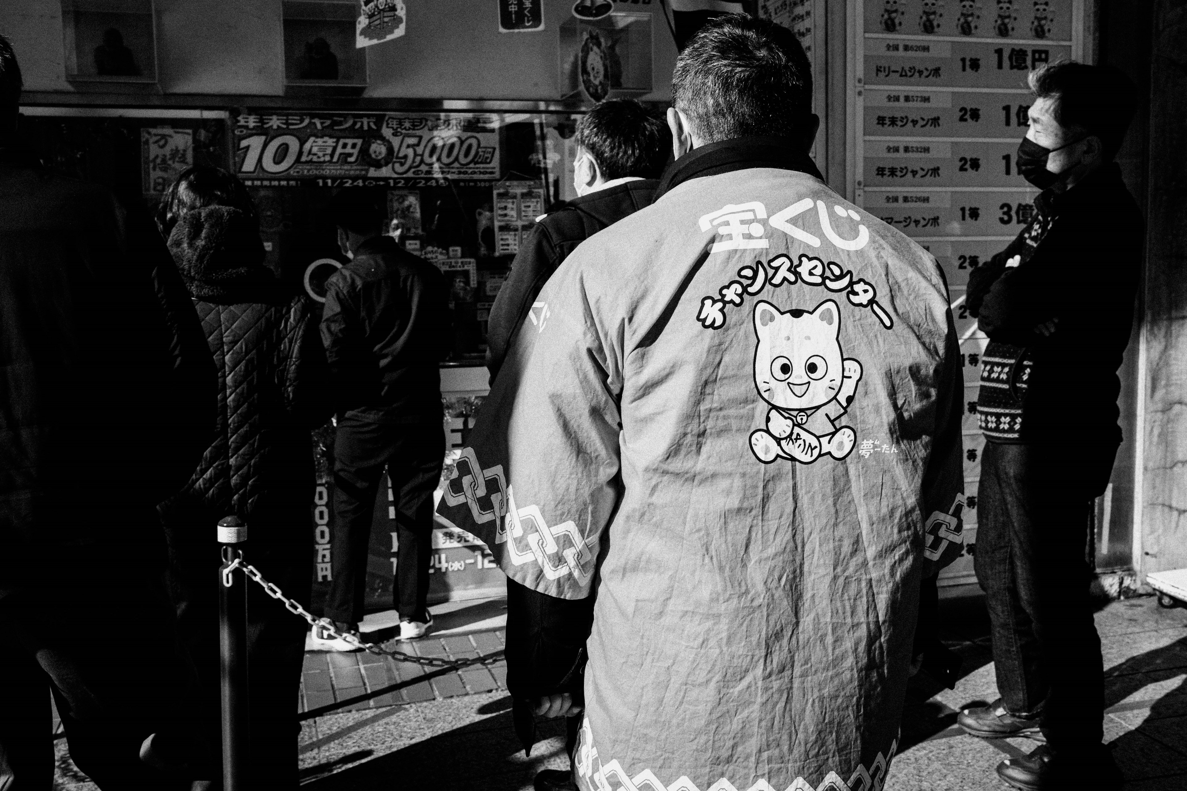 Image en noir et blanc montrant une file de personnes avec un homme portant un manteau à motif de personnage mignon au premier plan