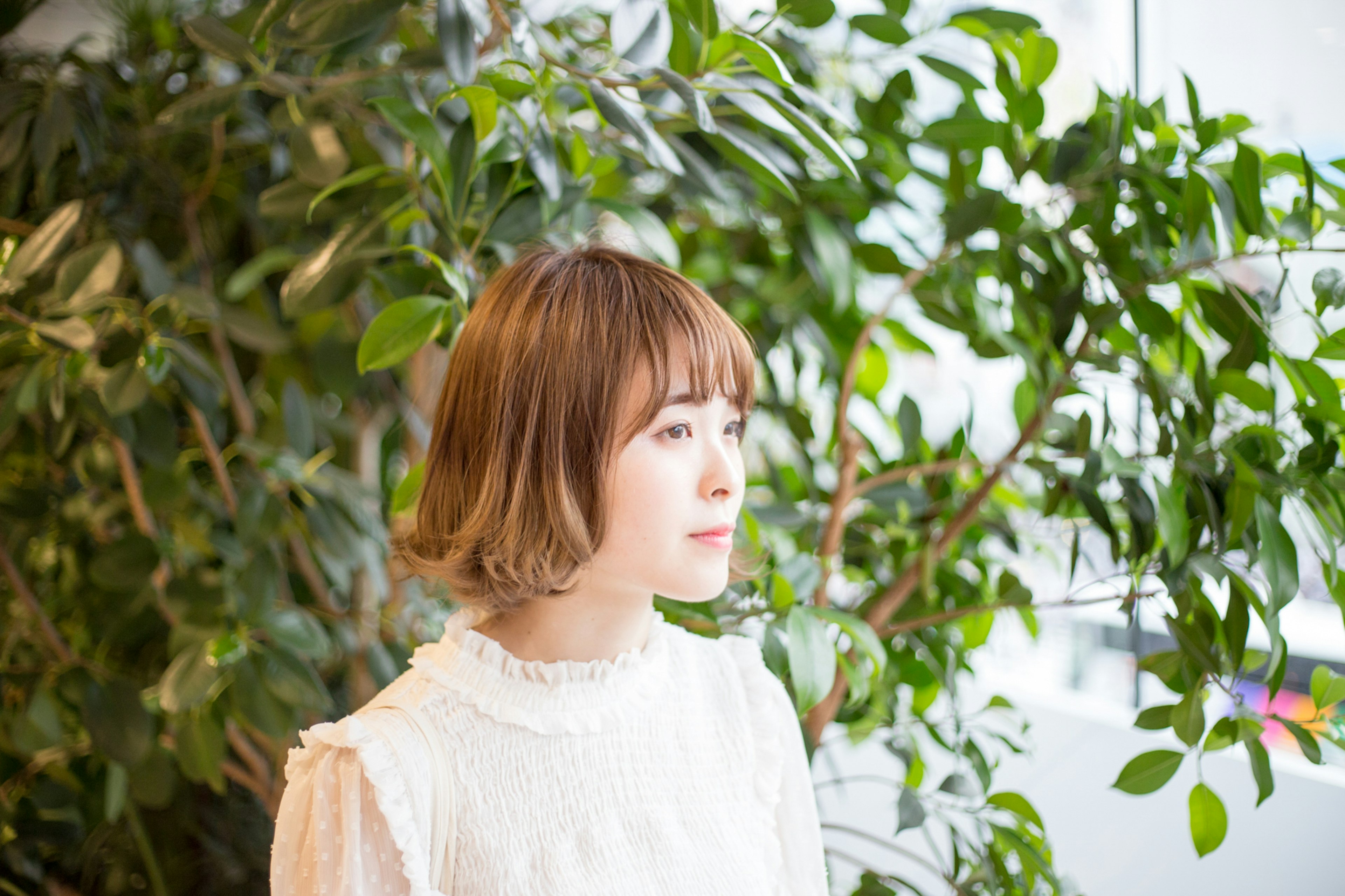 Portrait of a woman standing in front of green plants with a short hairstyle and wearing a white blouse