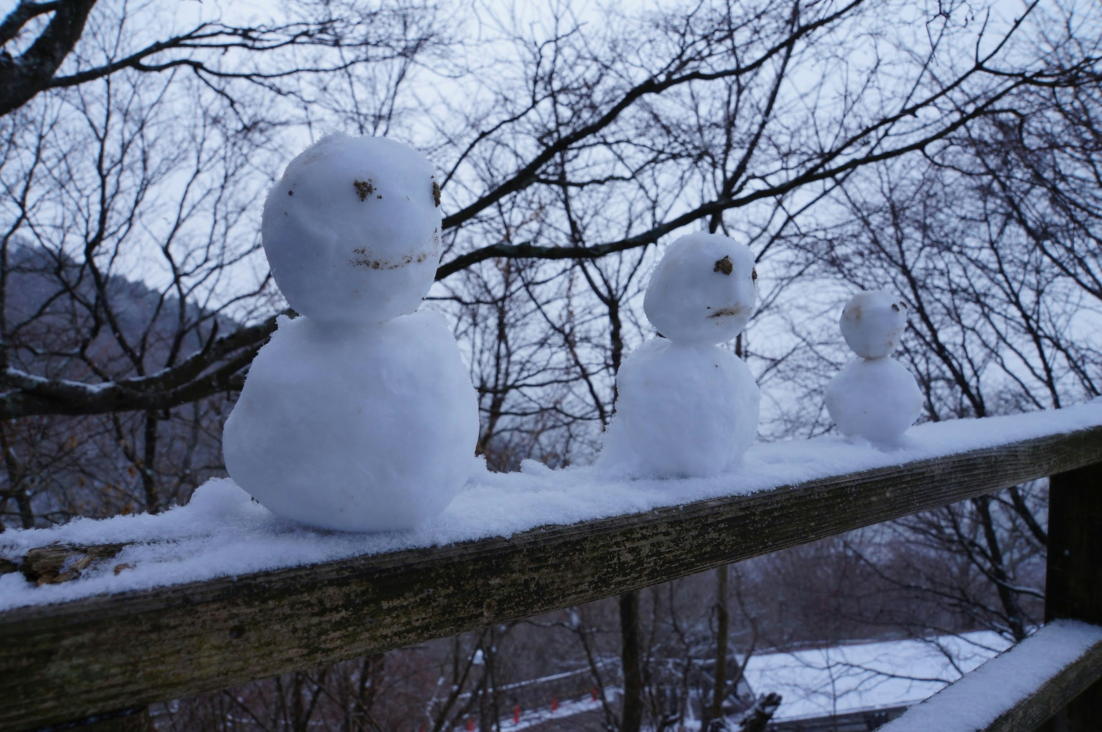 Tres muñecos de nieve sentados en una barandilla de madera rodeados de árboles