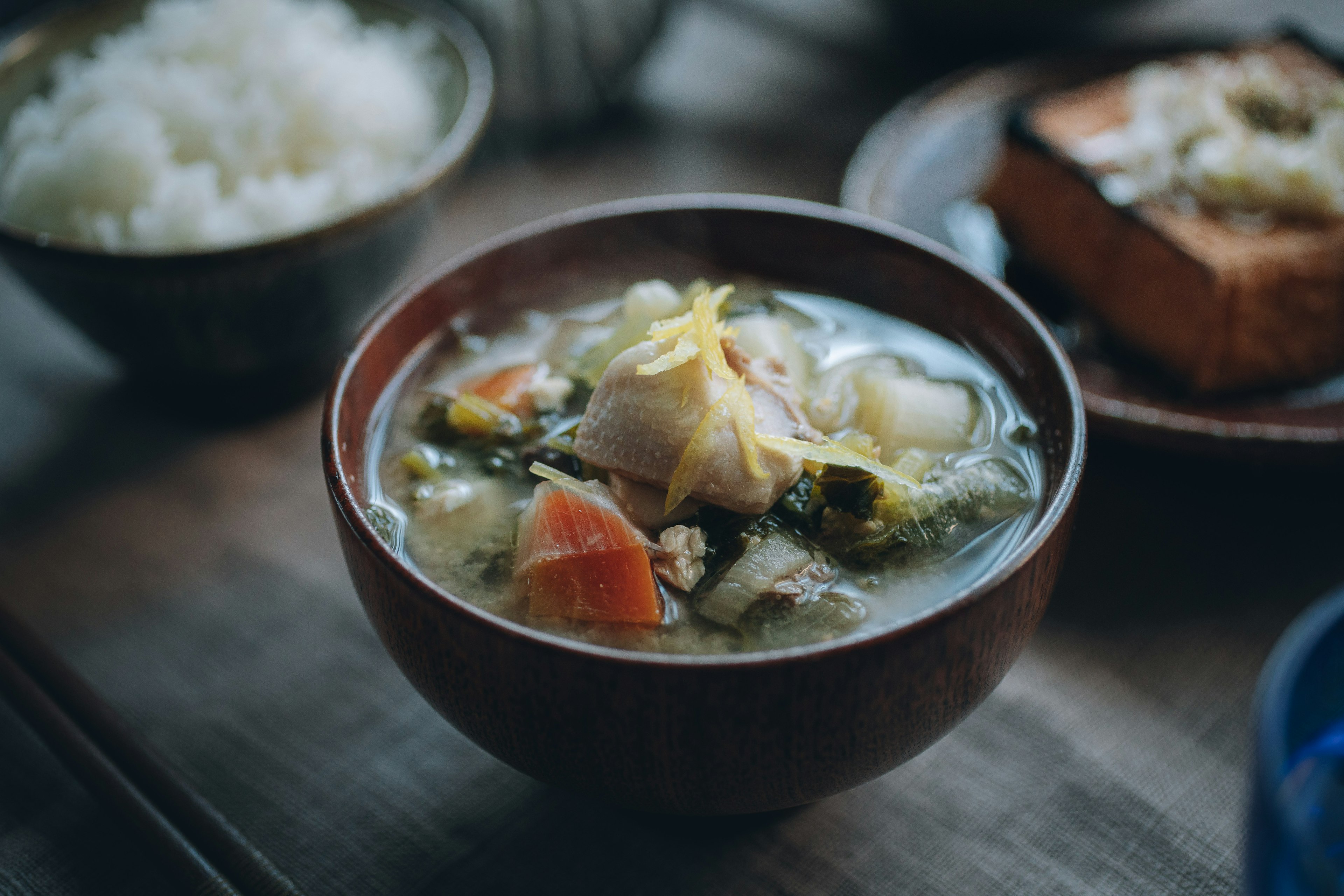 A bowl of warm soup with vegetables and a side of rice on a wooden table