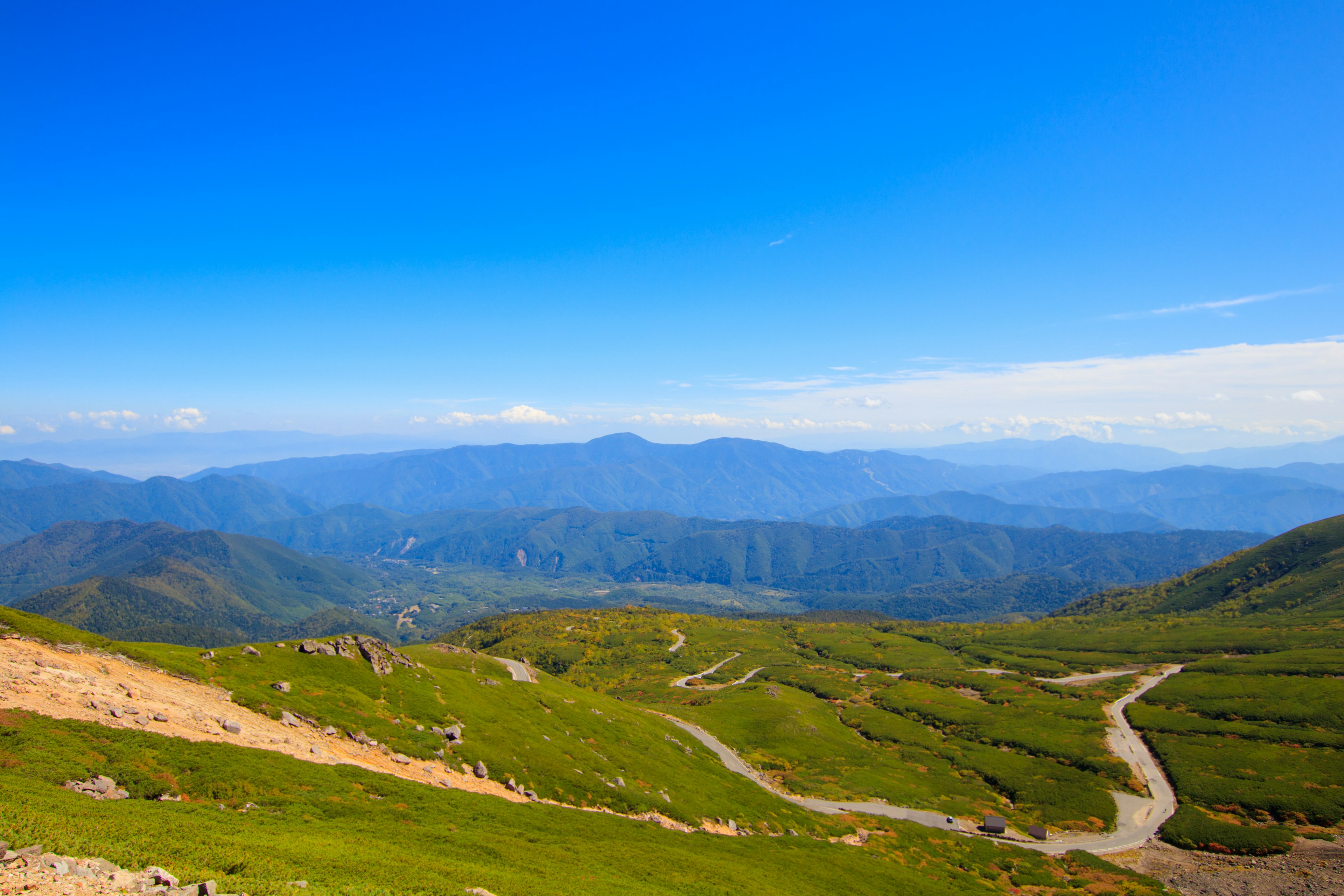 Vue panoramique de collines vertes avec des chemins sinueux sous un ciel bleu et des montagnes au loin