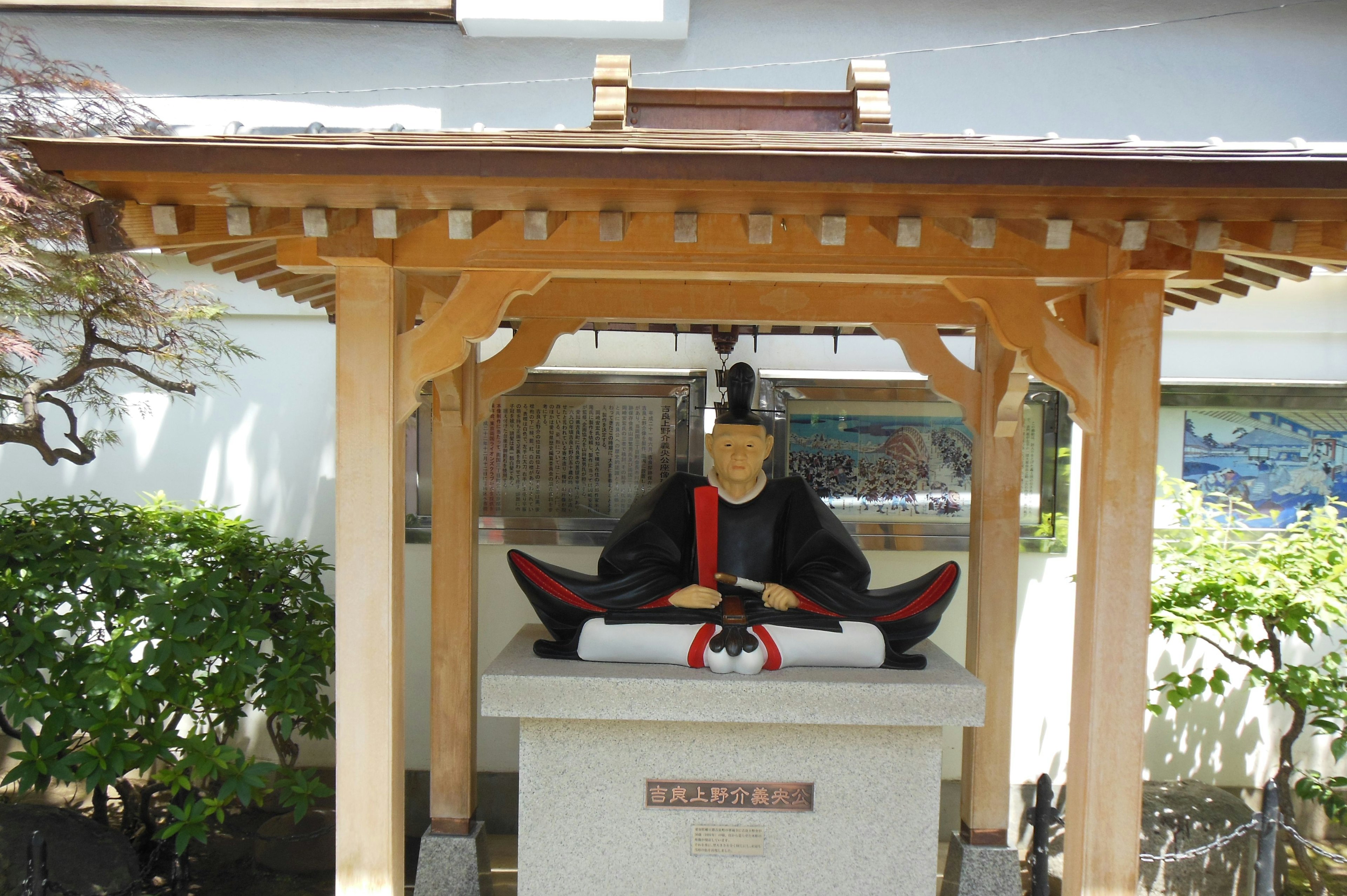 A statue of a samurai sitting under a wooden roof with green plants surrounding it