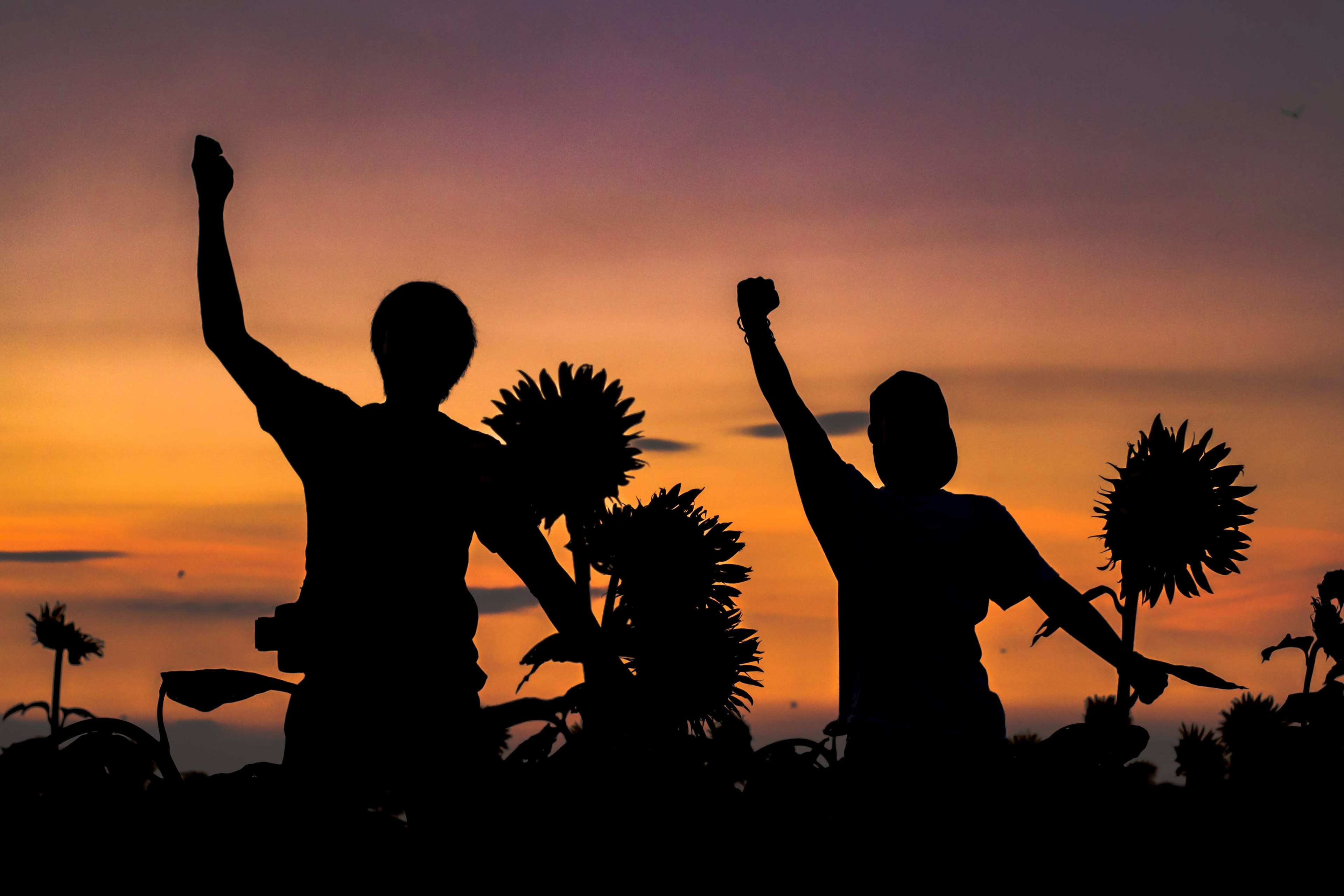 Silueta de dos personas levantando las manos contra un atardecer con girasoles