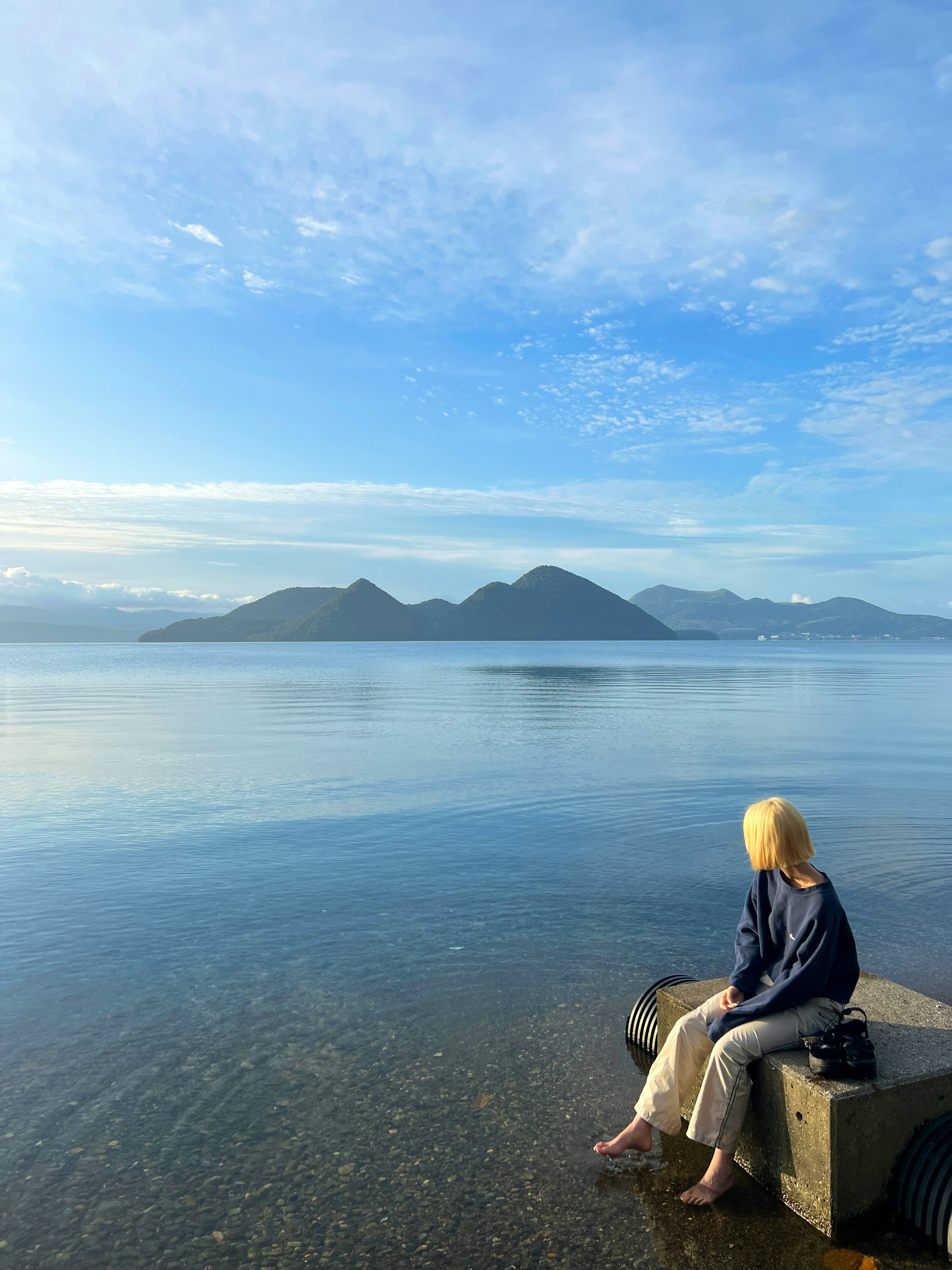 Mujer sentada en una piedra junto a un lago tranquilo bajo un cielo azul