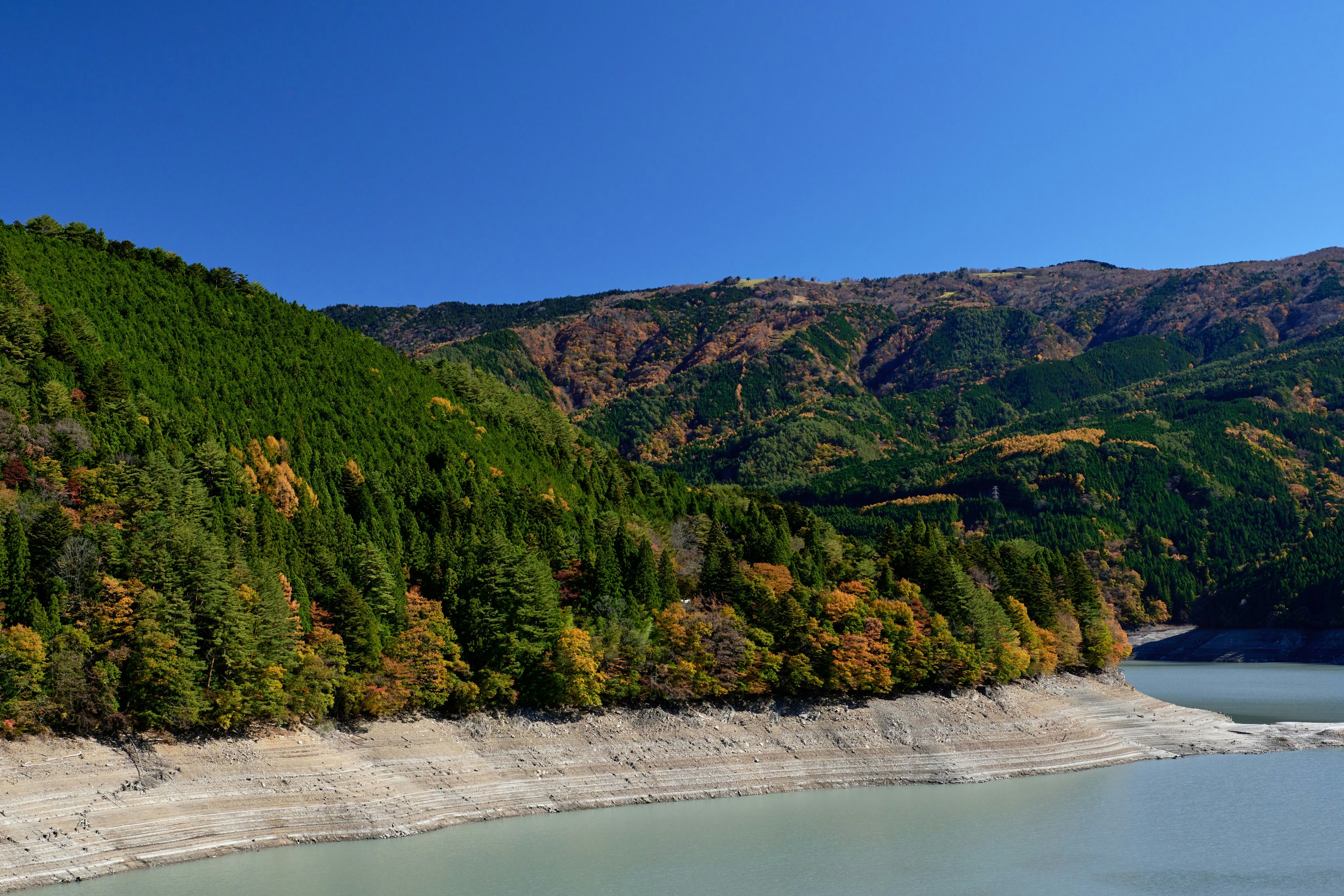 Landschaftsansicht eines Sees mit grünen Bergen und Herbstlaub unter blauem Himmel