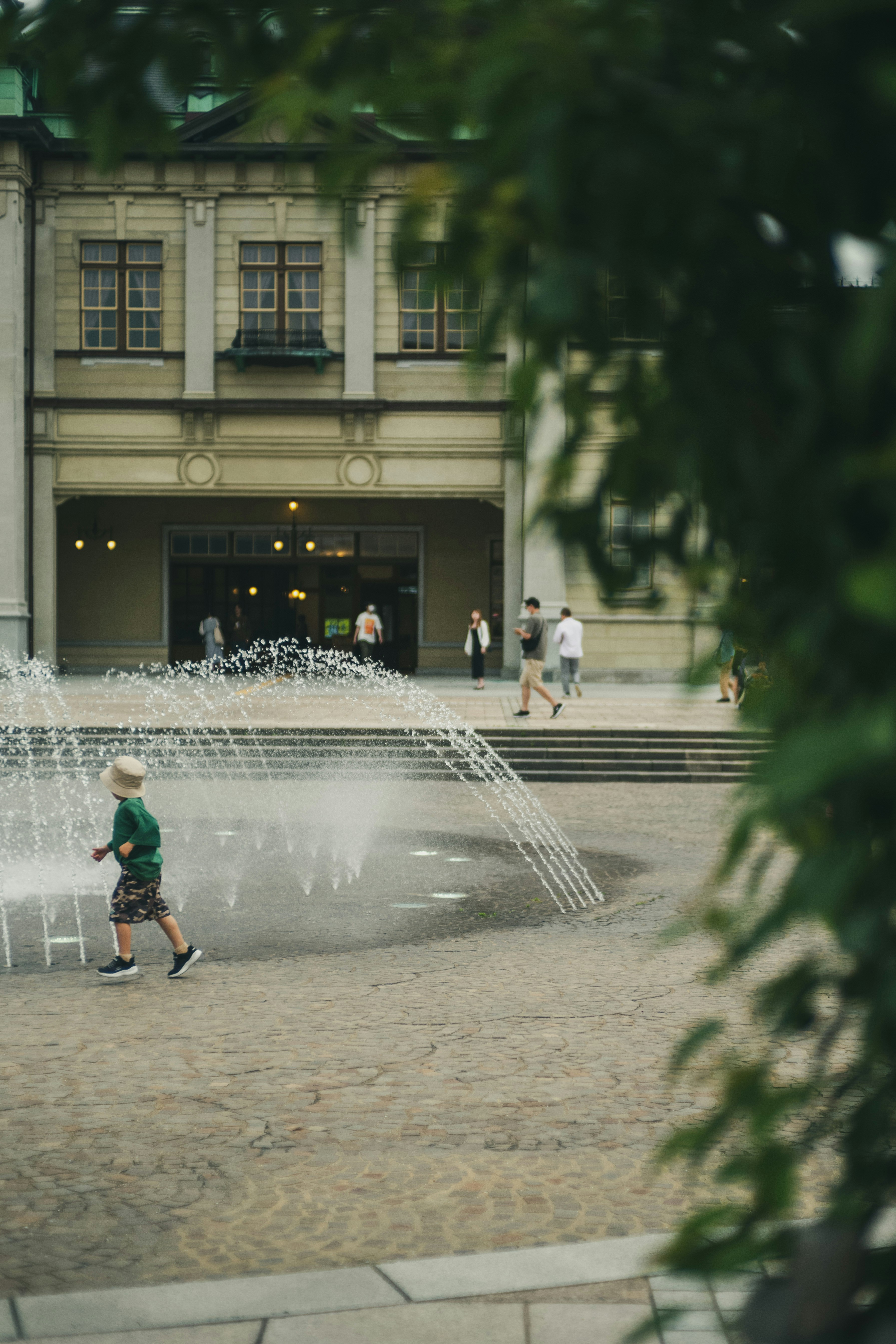 A child in a green shirt running near a fountain in an urban setting