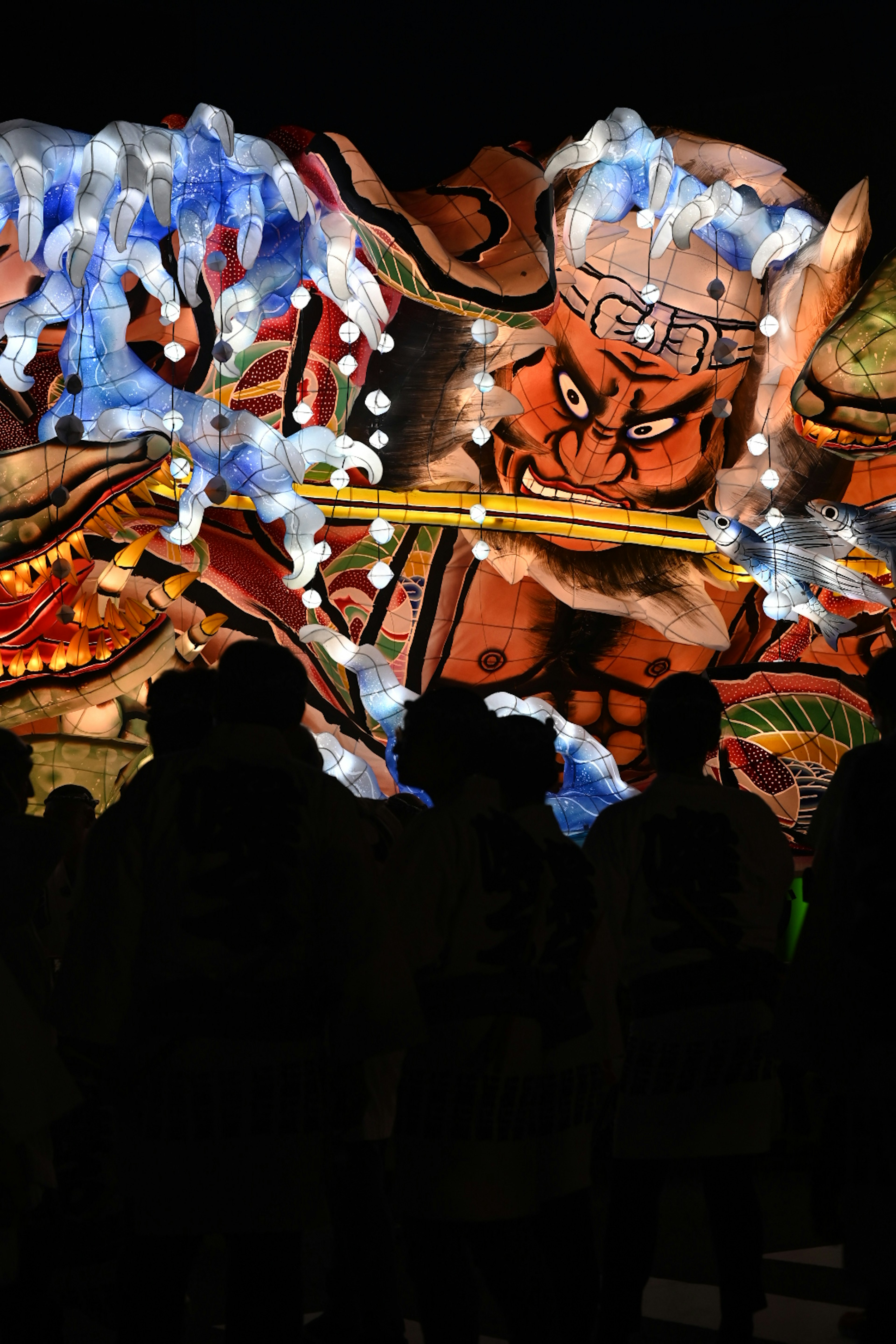 People standing in front of a giant illuminated lantern depicting a fierce demon at night festival