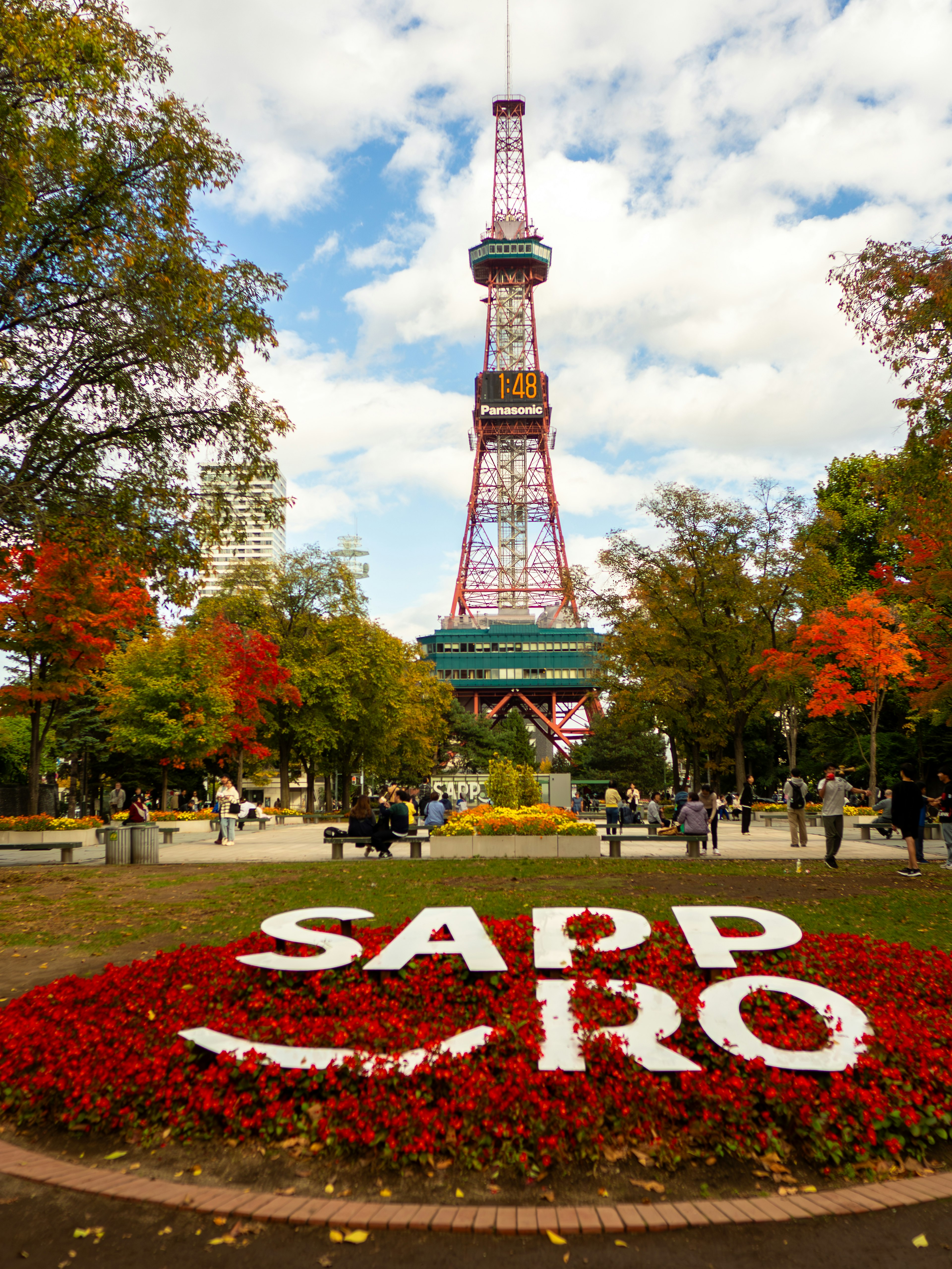 Pemandangan Menara TV Sapporo di taman dengan pepohonan musim gugur berwarna-warni dan taman bunga yang mengeja 'SAPPORO'