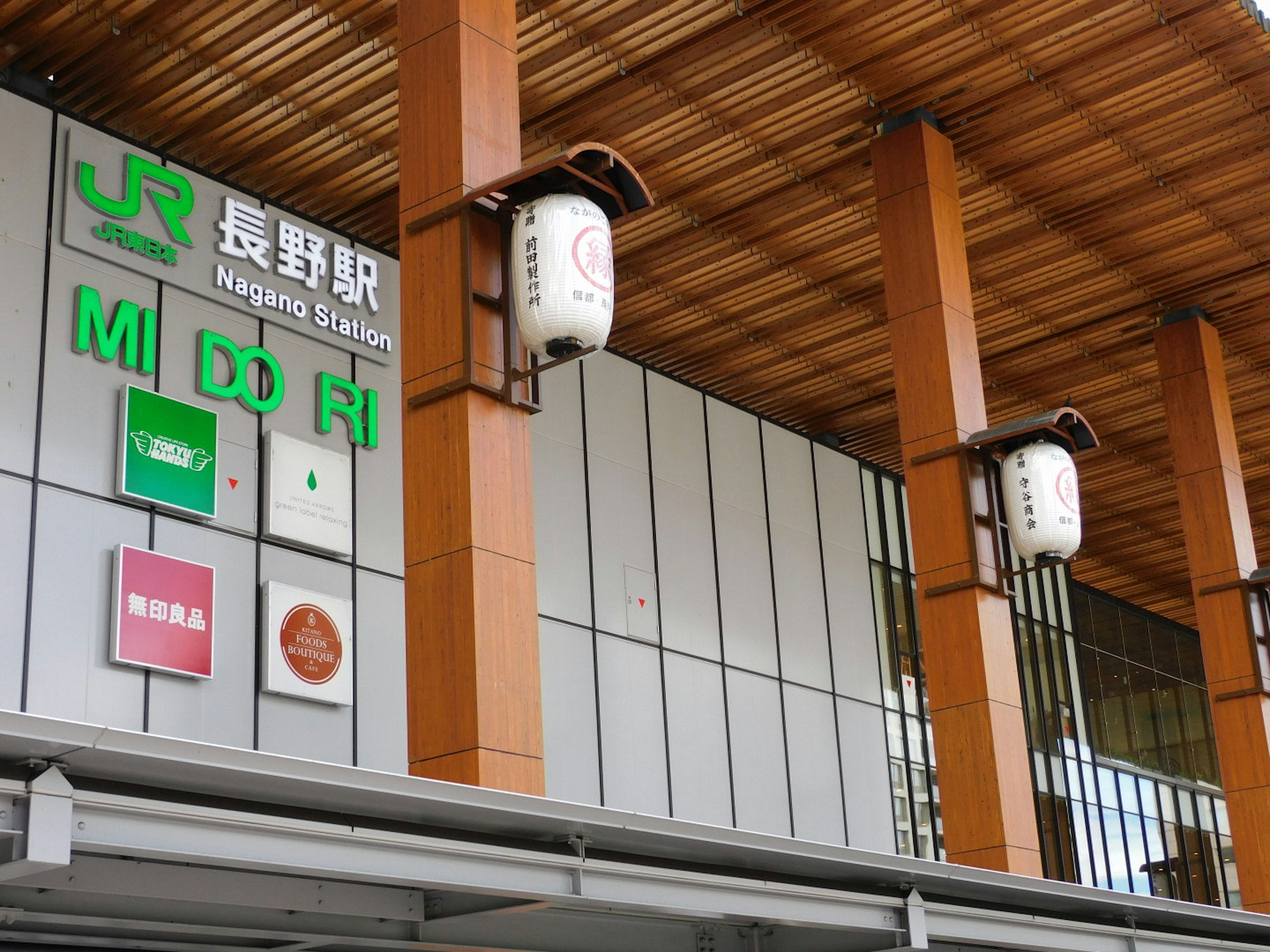 Außenansicht des Nagano Bahnhofs mit Beschilderung in Japanisch und Englisch sowie dem Bahnhofslogo