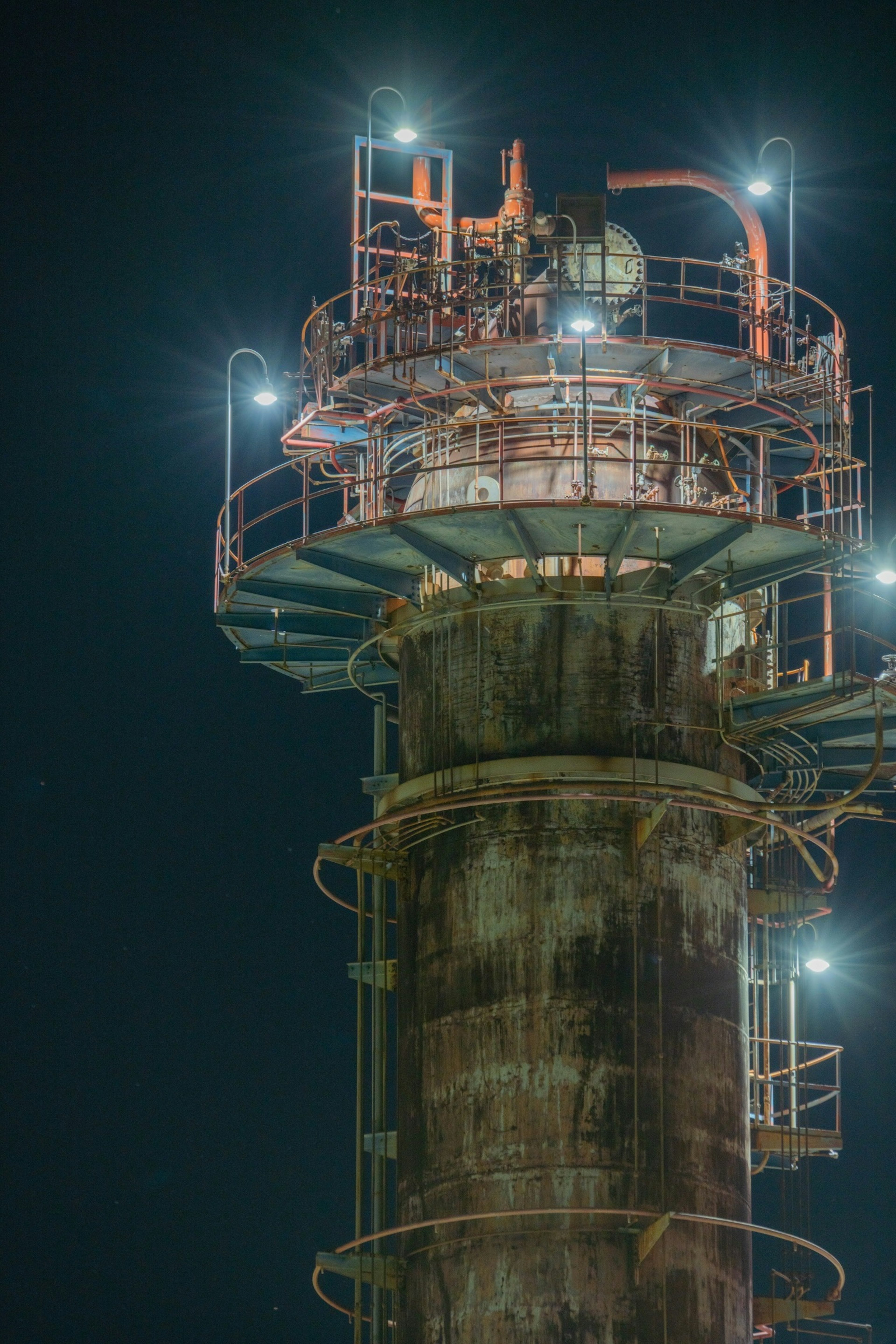 Detailed image of an industrial tower at night featuring rusted metal and bright lights