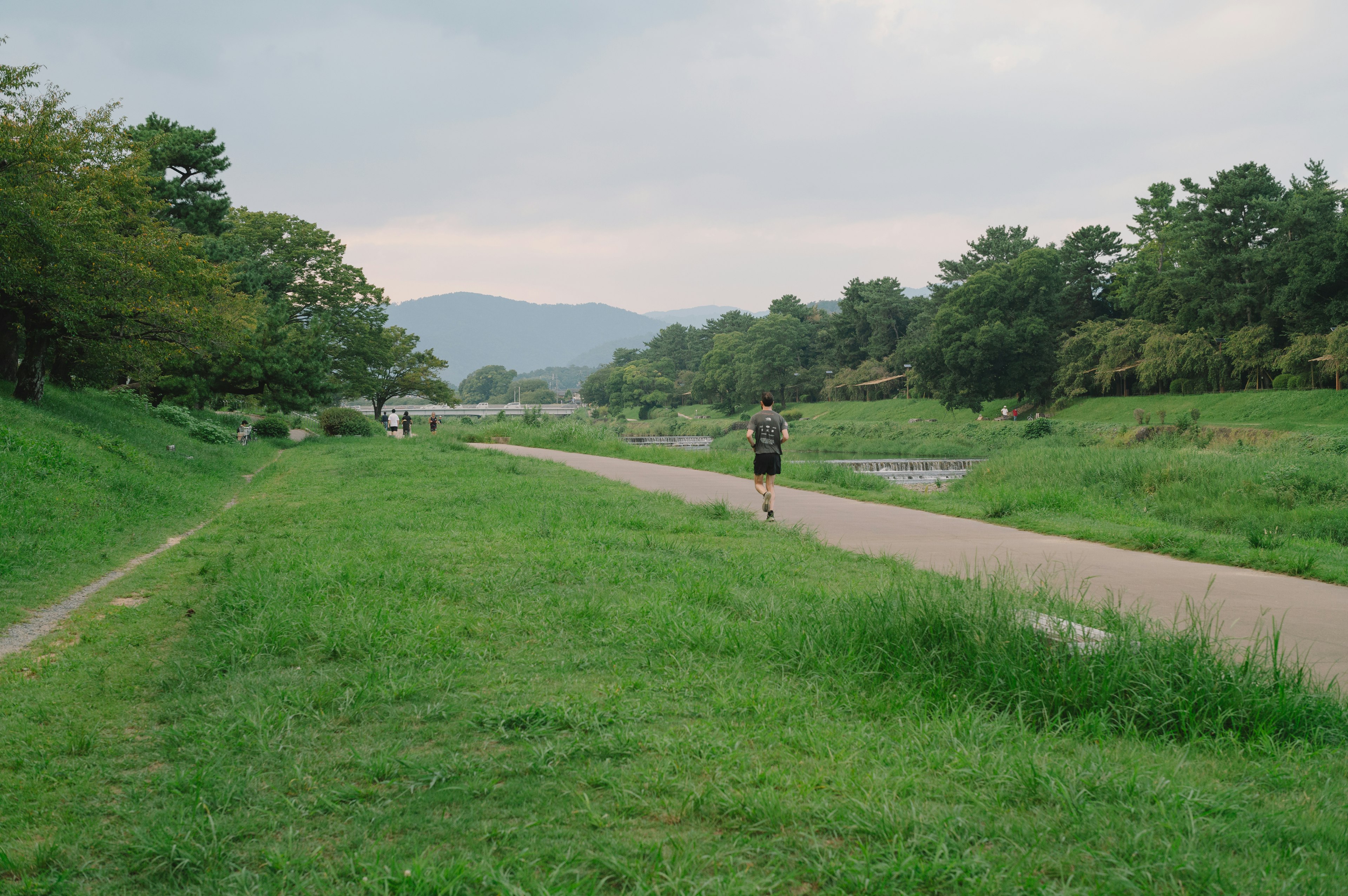 Un hombre corriendo por un camino de hierba junto a un río con árboles y montañas al fondo