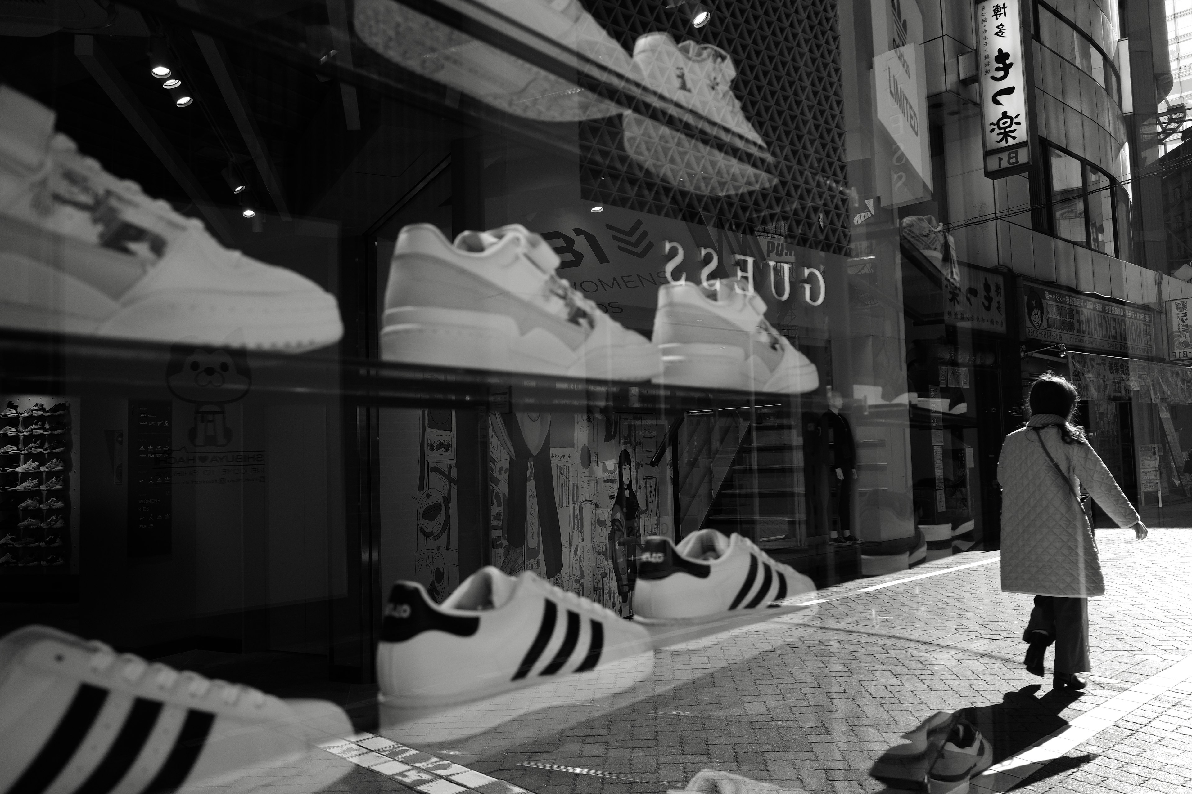 Photo en noir et blanc présentant des baskets Adidas dans une vitrine avec une personne marchant