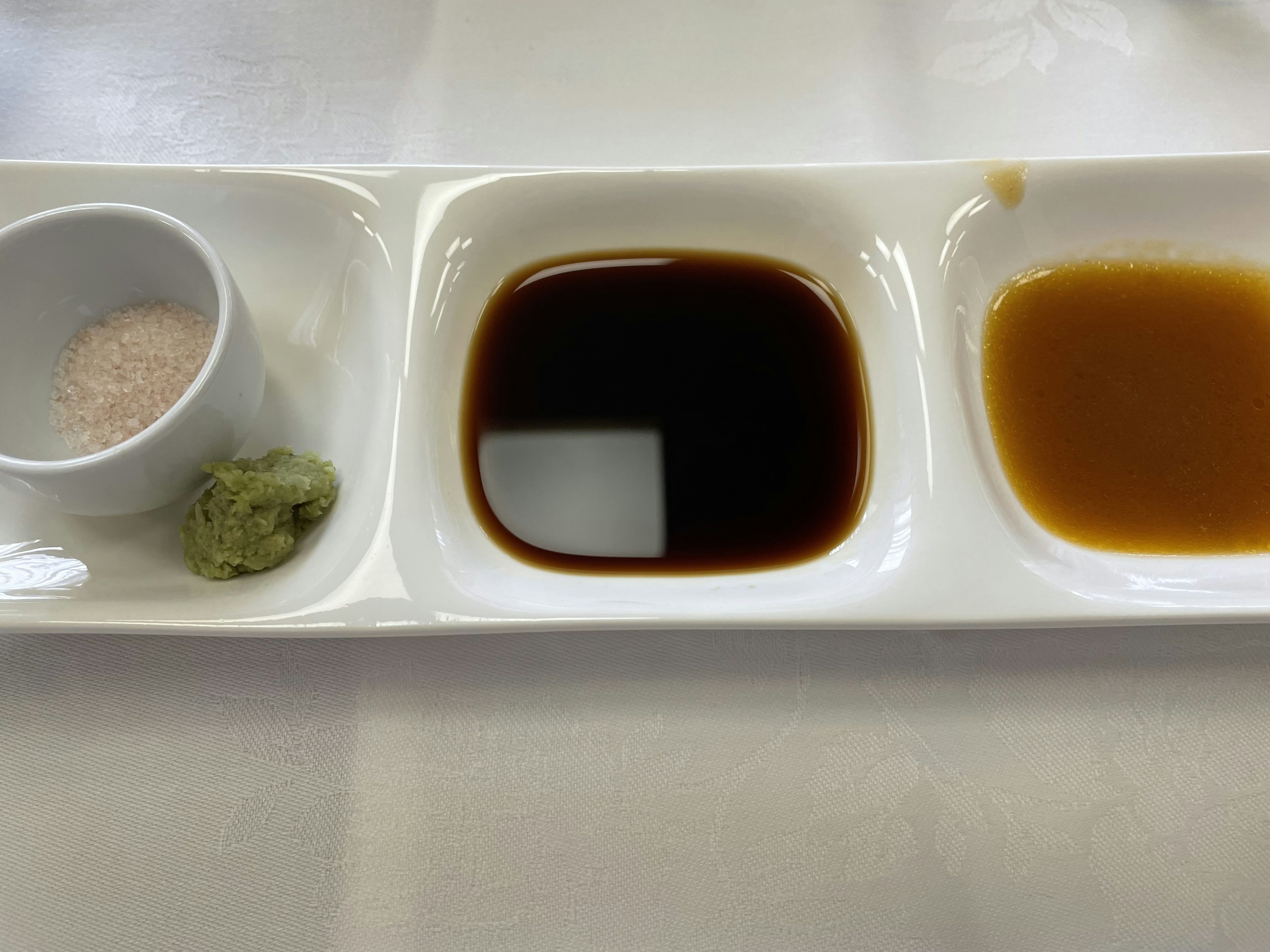 Tres condimentos dispuestos en un plato blanco que incluyen salsa de soja, miso, sal y wasabi