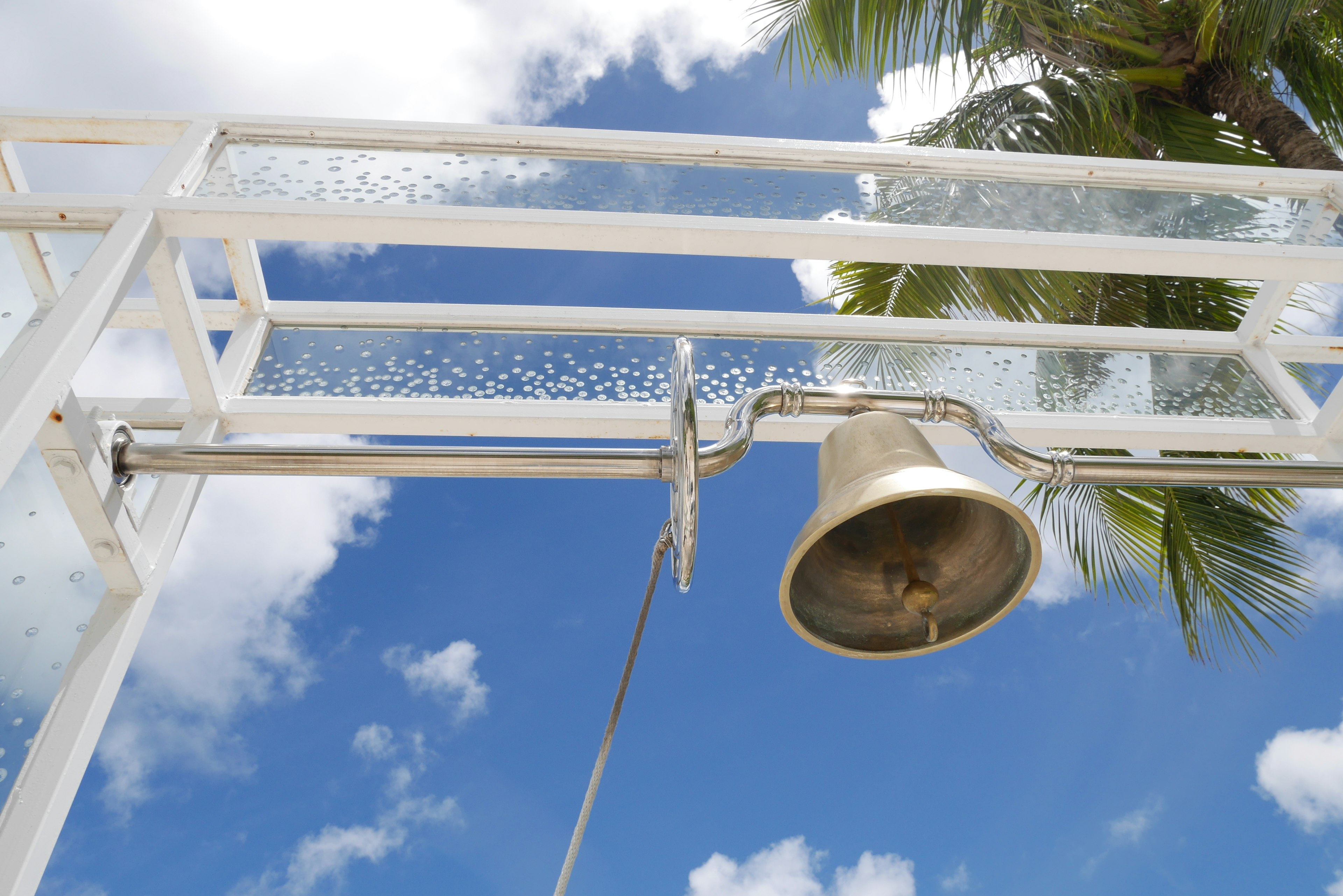 Bell hanging from a white frame under a blue sky with palm trees