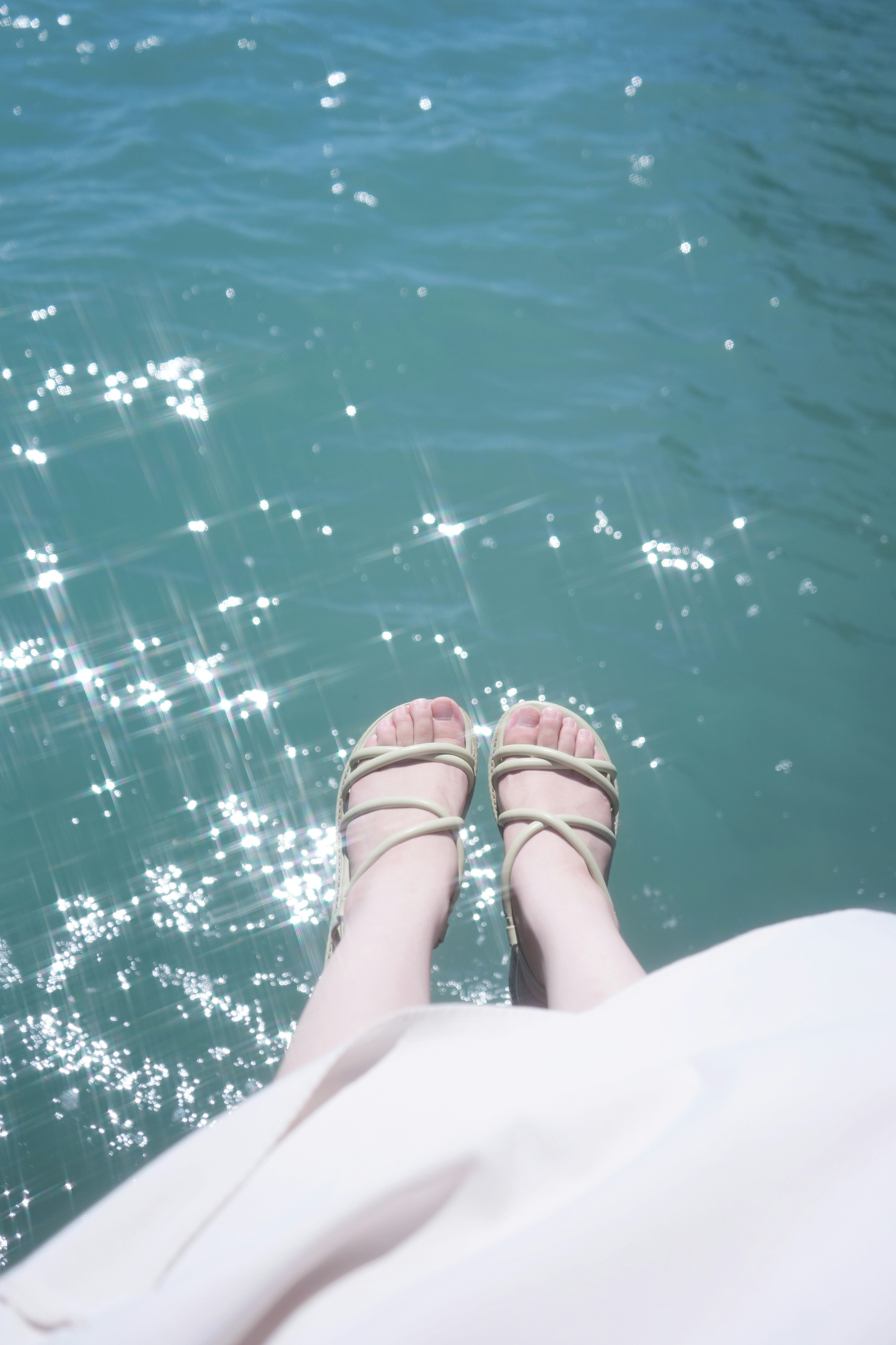 A person's feet in sandals dangling over sparkling blue water