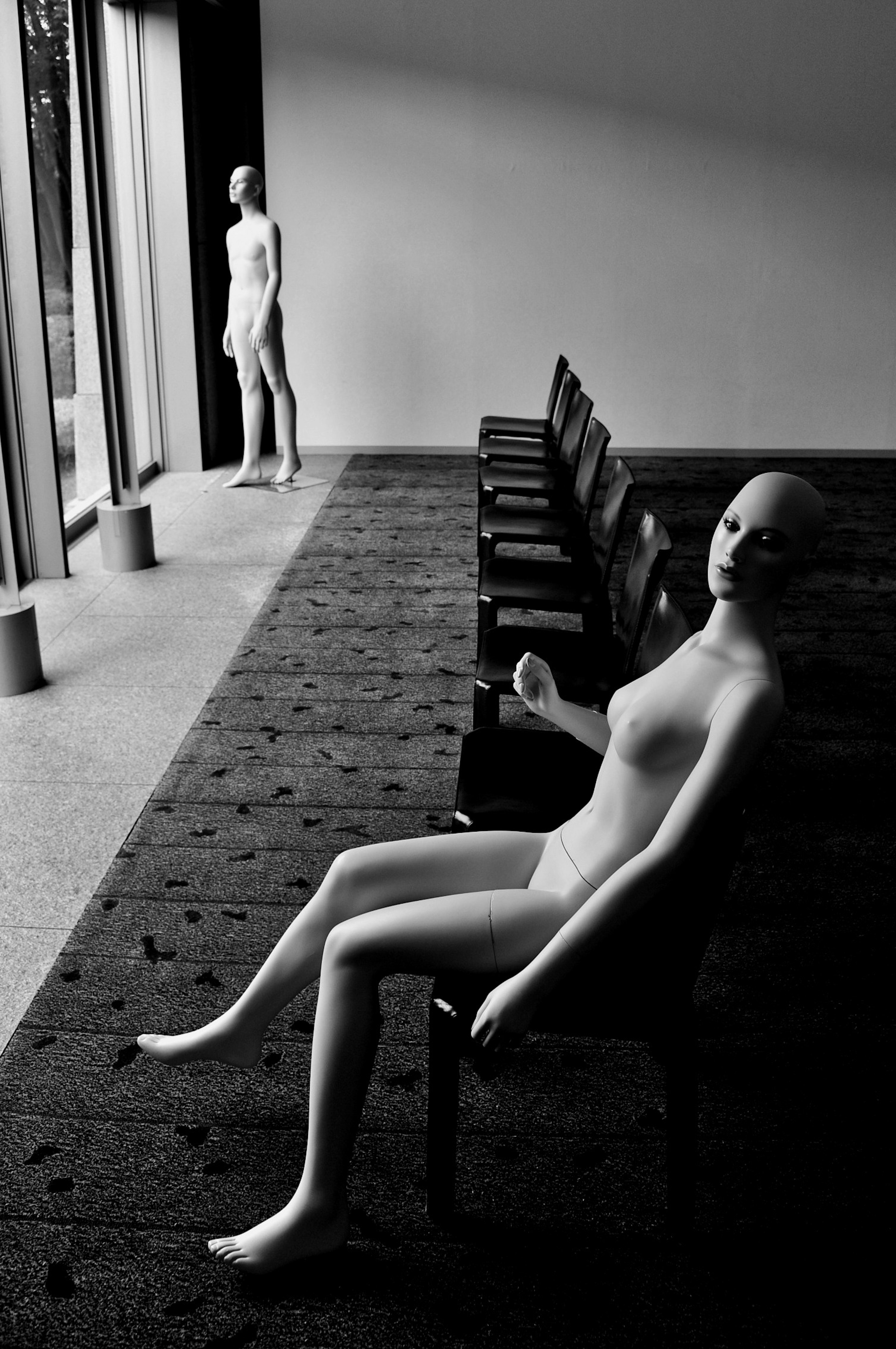 Una imagen en blanco y negro de un maniquí sentado en una silla con otro maniquí de pie al fondo