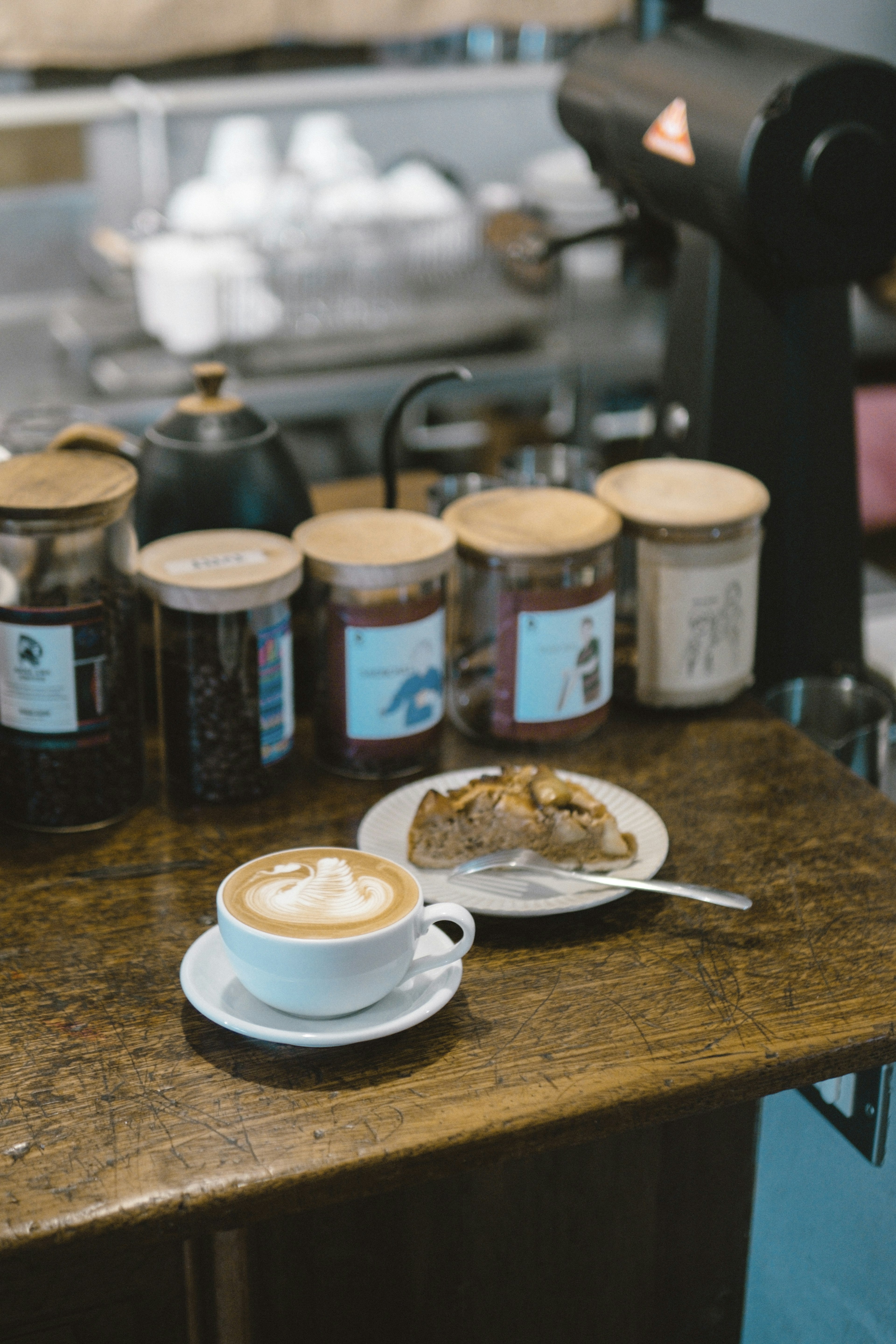 A coffee cup with latte art and a slice of cake on a plate on a wooden table with jars of coffee beans in the background