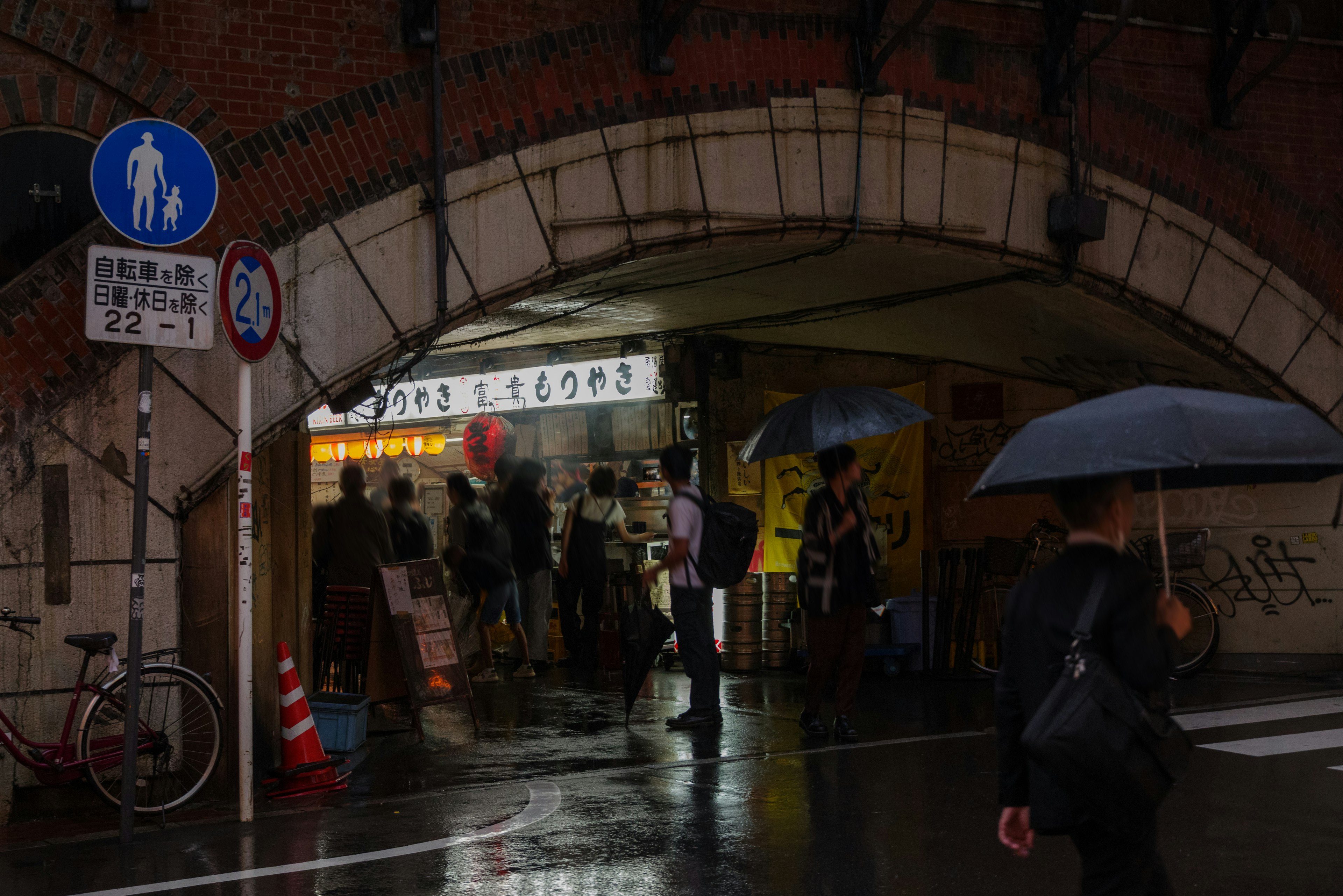 Persone con ombrelli che camminano sotto un arco di mattoni con insegne e bancarelle di cibo visibili