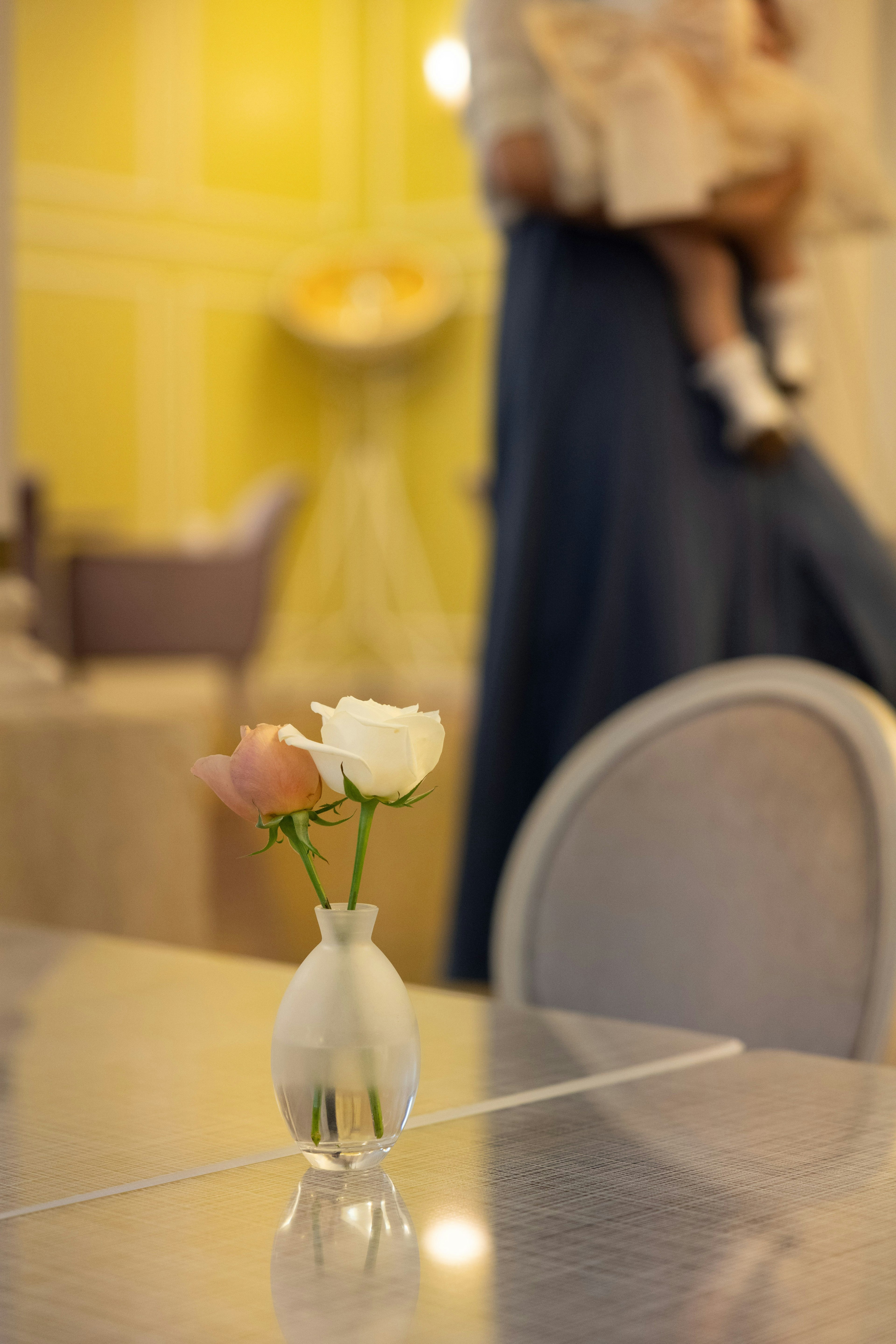 A small vase on a table with pink and white flowers in it A person holding a child is blurred in the background