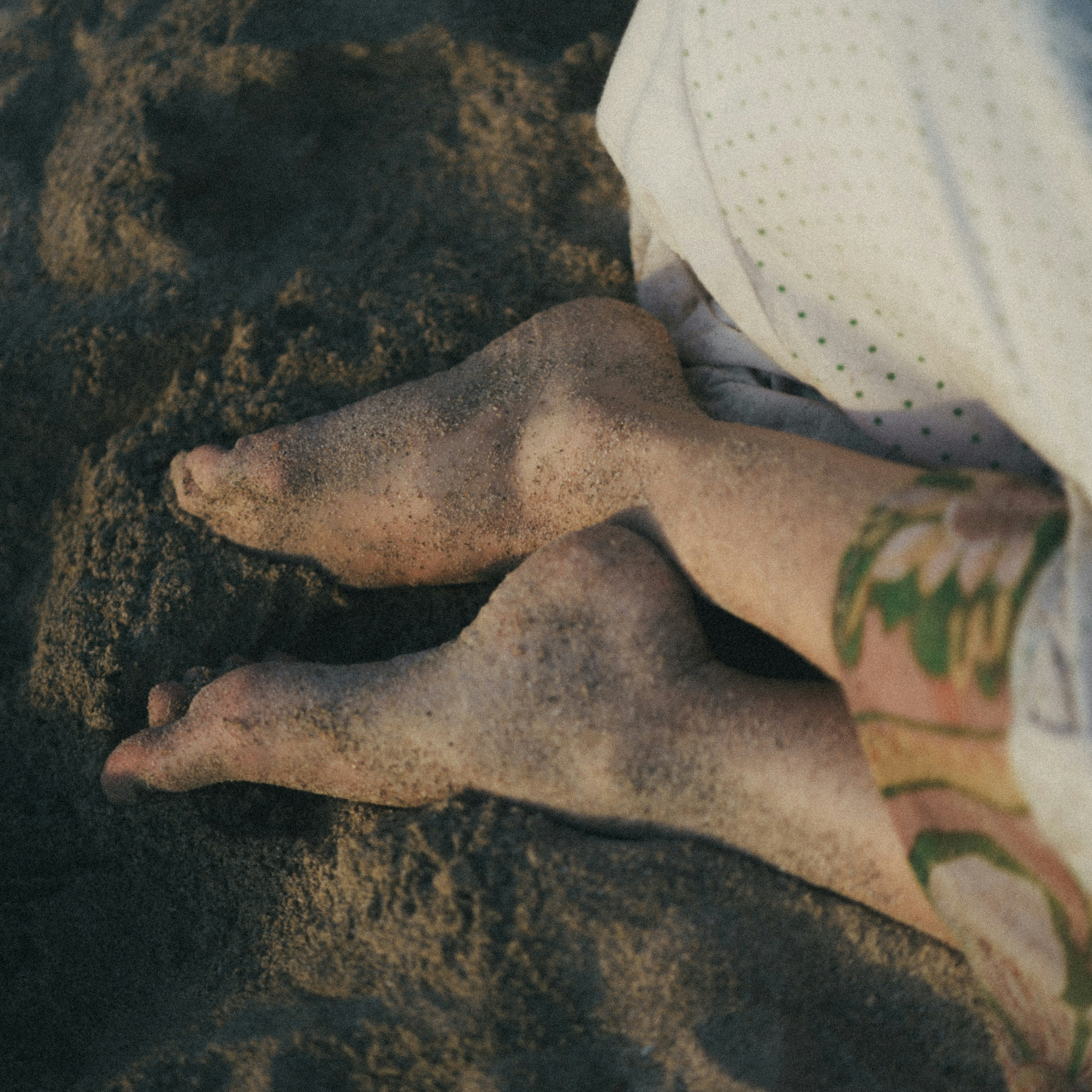 Füße einer Person mit sichtbarem Tattoo, die am Sandstrand sitzt