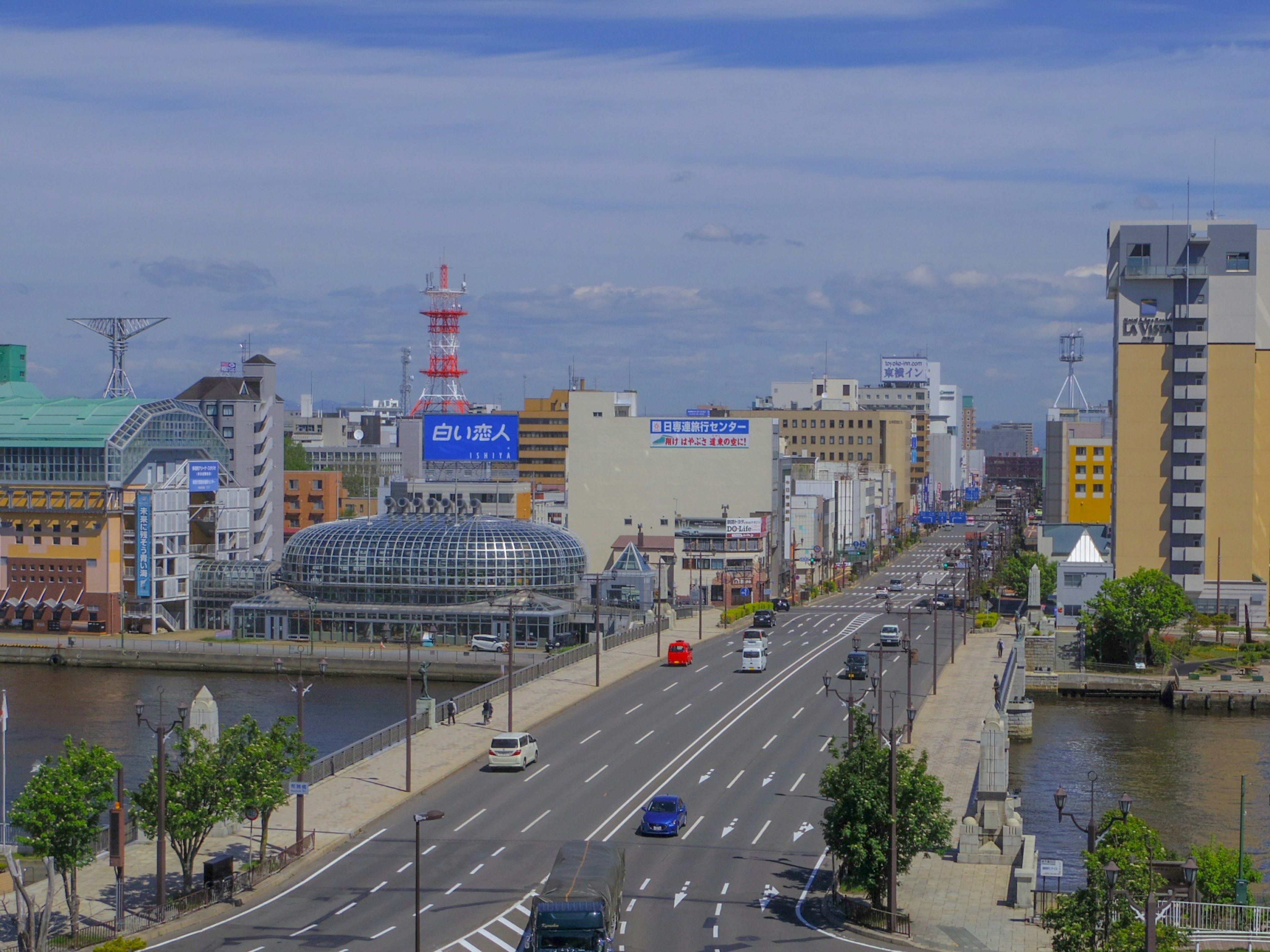 Cityscape featuring a wide road along a river, buildings, and a clear blue sky