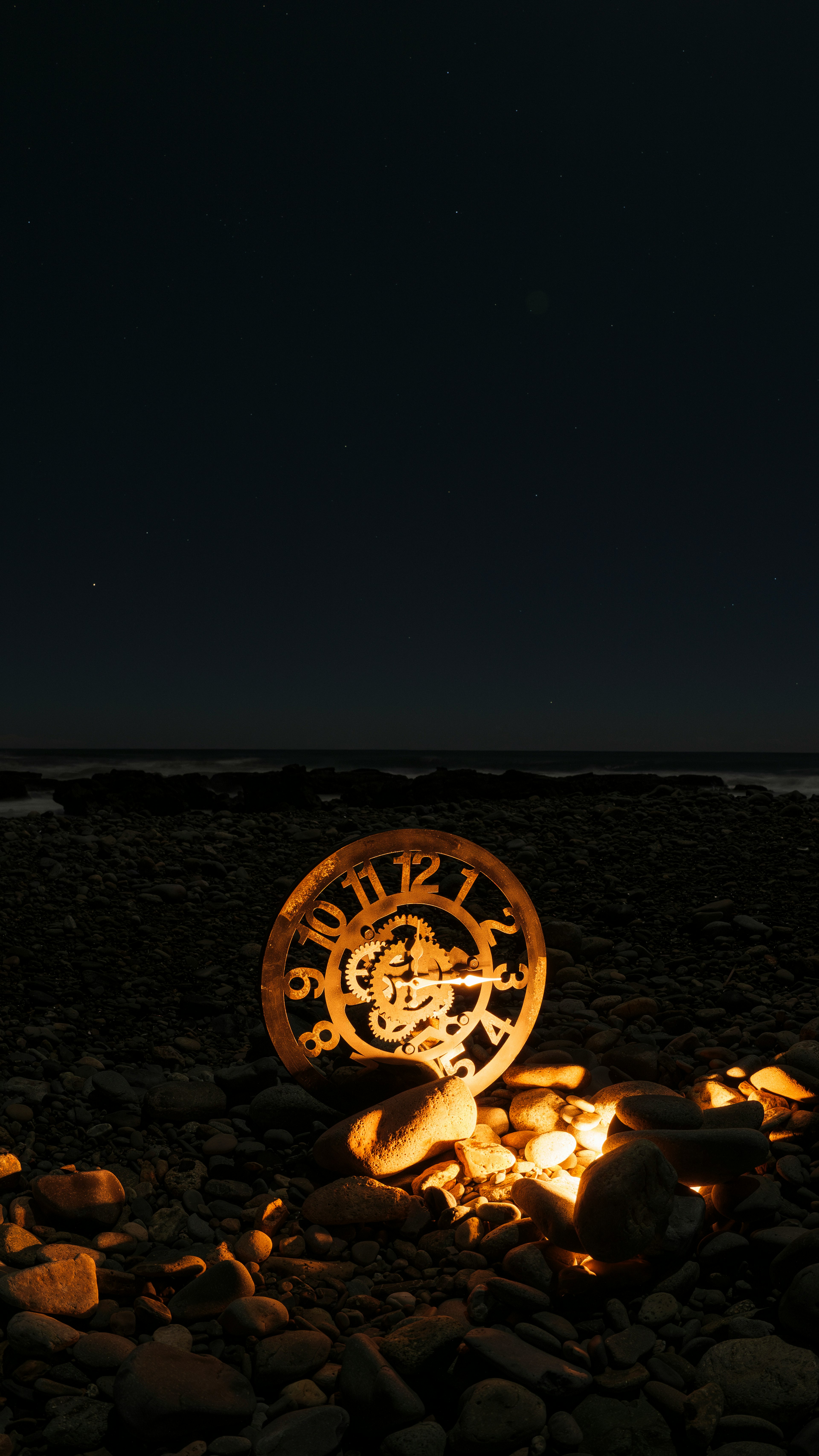 An old clock surrounded by stones on a beach at night