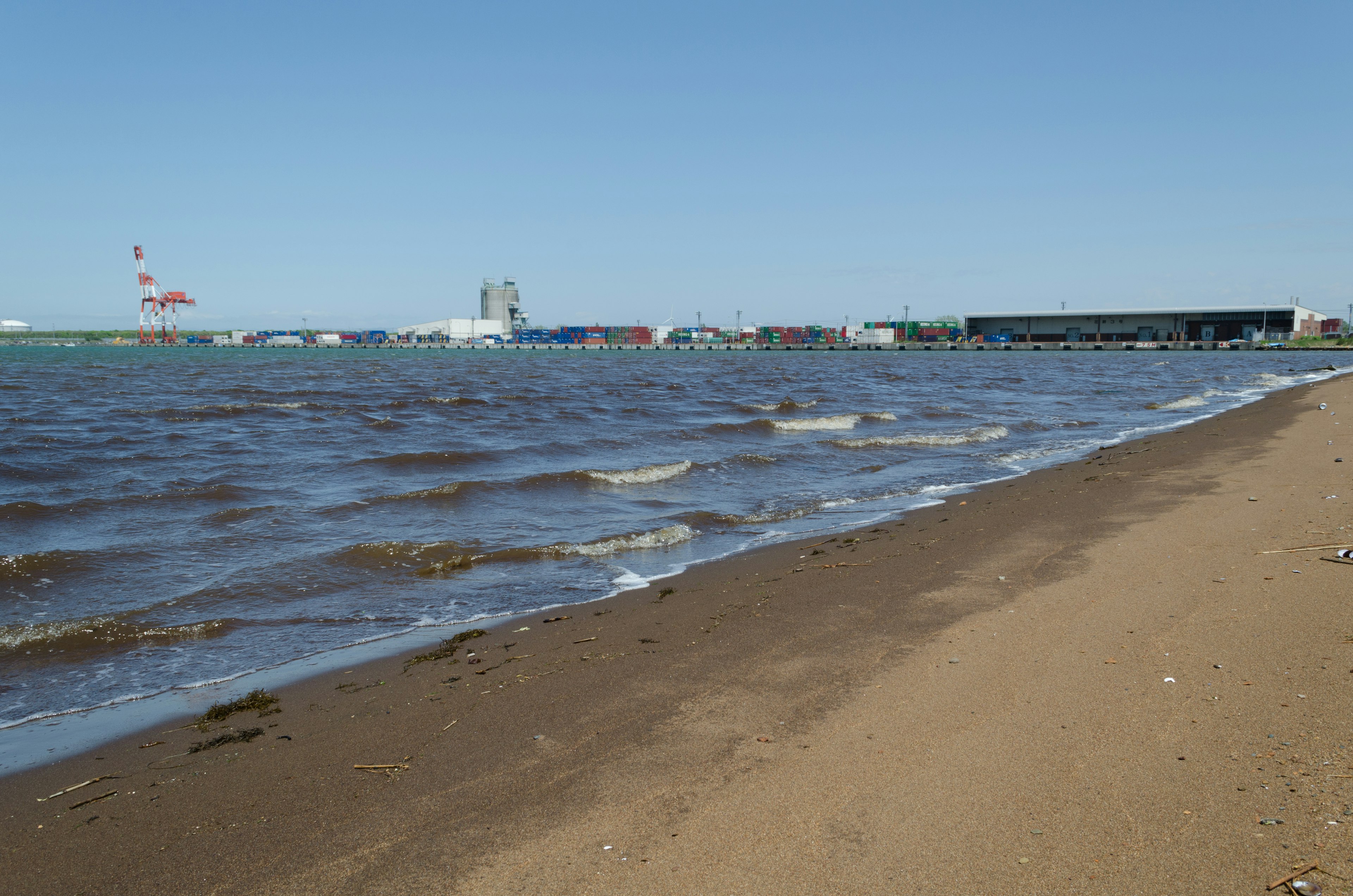 Pantai berpasir dengan ombak lembut dan bangunan industri di kejauhan