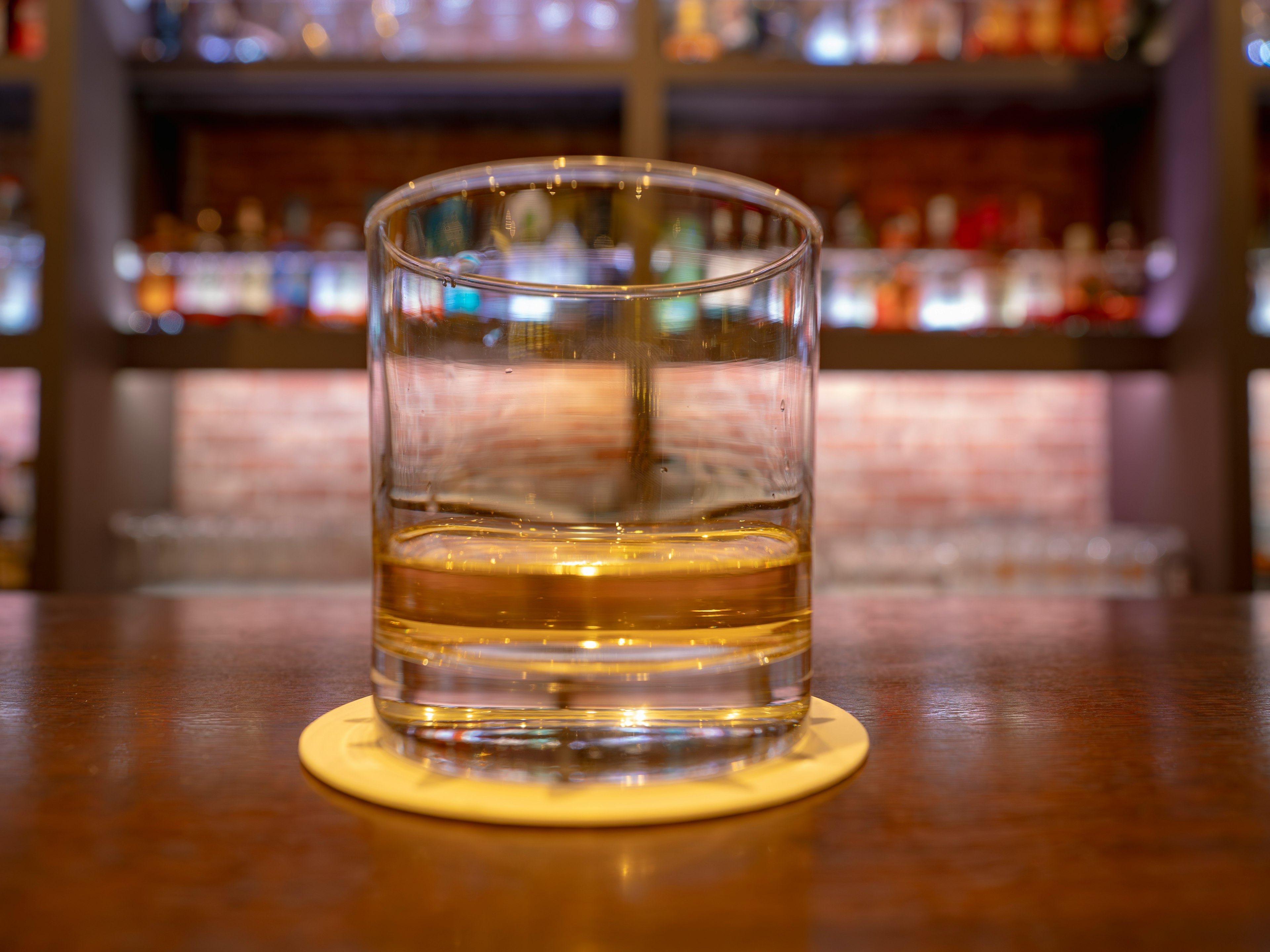 Whiskey dalam gelas bening di atas tatakan dengan latar belakang bar