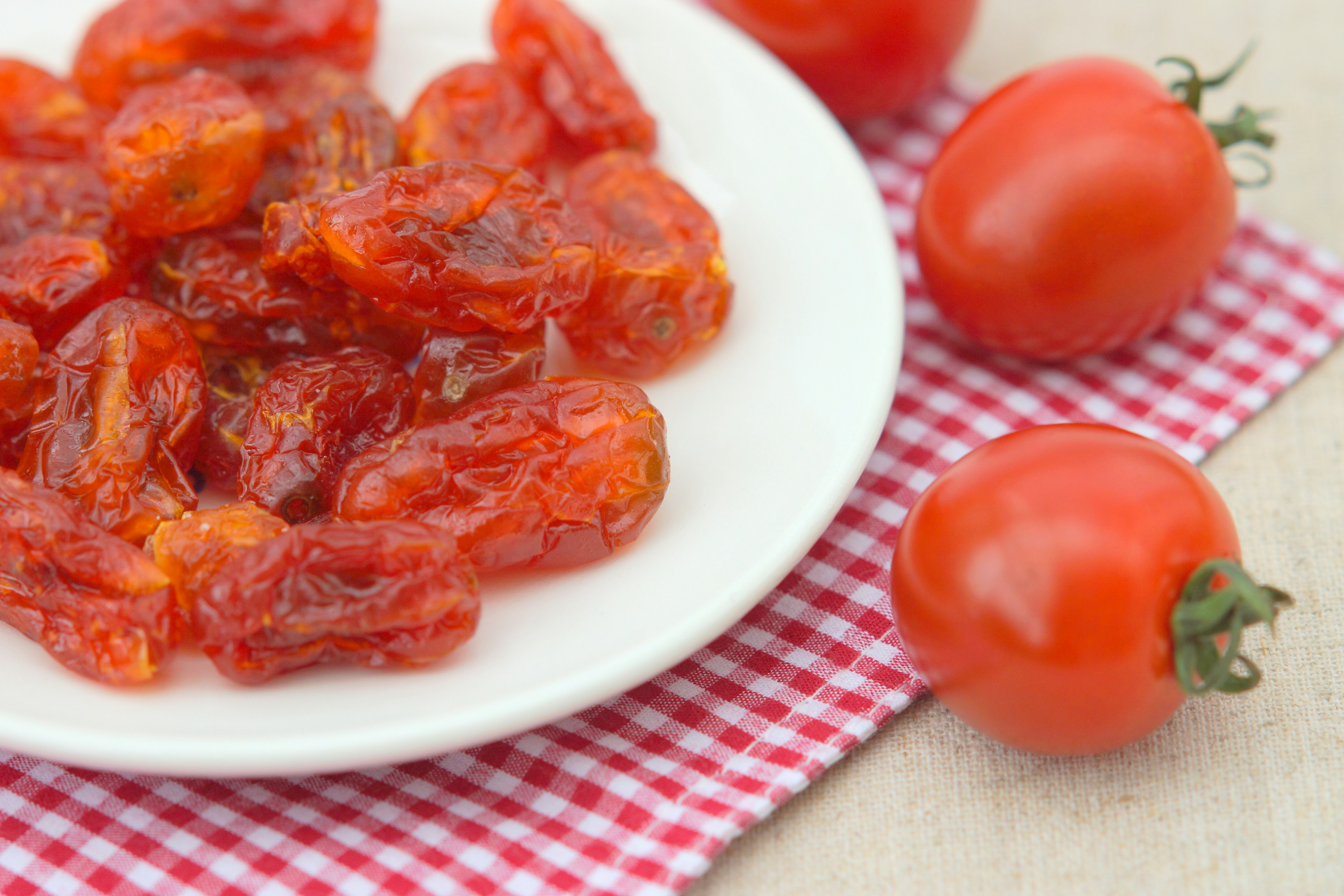 Tomates secas en un plato blanco con tomates frescos al lado sobre una tela a cuadros rojos